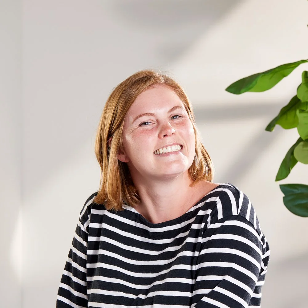 Smiling woman with shoulder-length red hair wearing a black and white striped shirt, sitting indoors near a green plant.