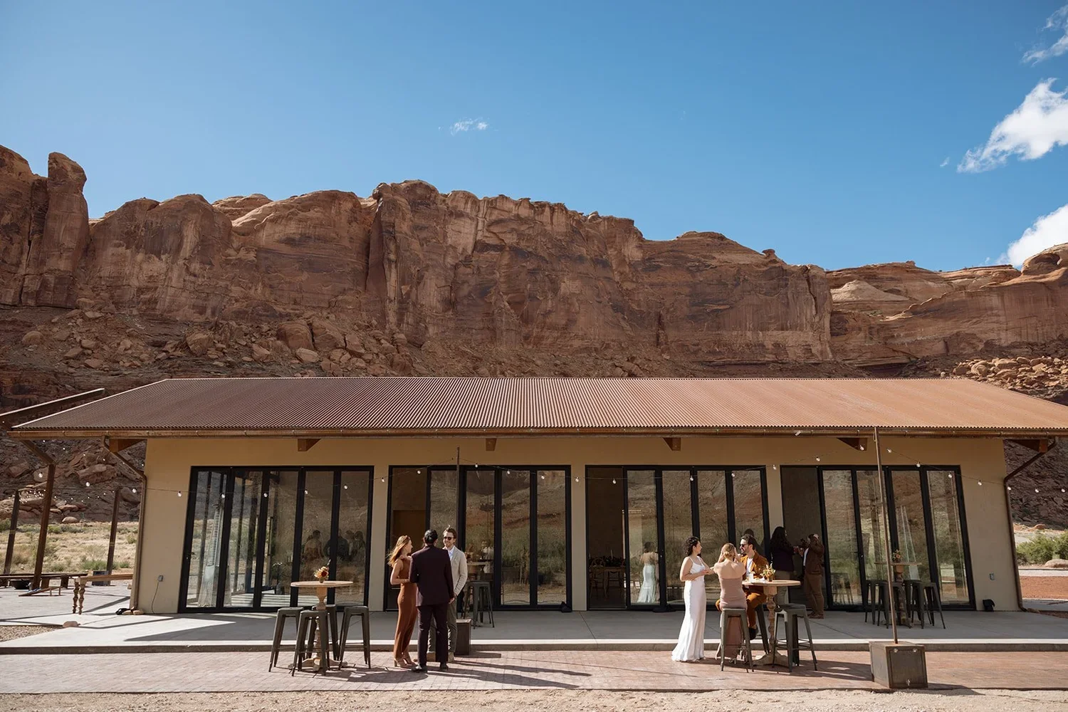 Group of people socializing outside a modern building with large glass windows, set against a desert landscape with red rock cliffs. Some are dressed in formal or semi-formal attire, and the scene appears to be at a gathering or celebration.