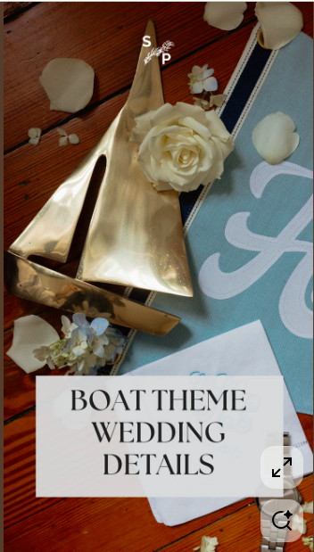 A gold-colored boat-shaped wedding cake topper with white flowers, placed on a table decorated for a wedding with scattered flower petals and a blue table runner that says 'Love'.