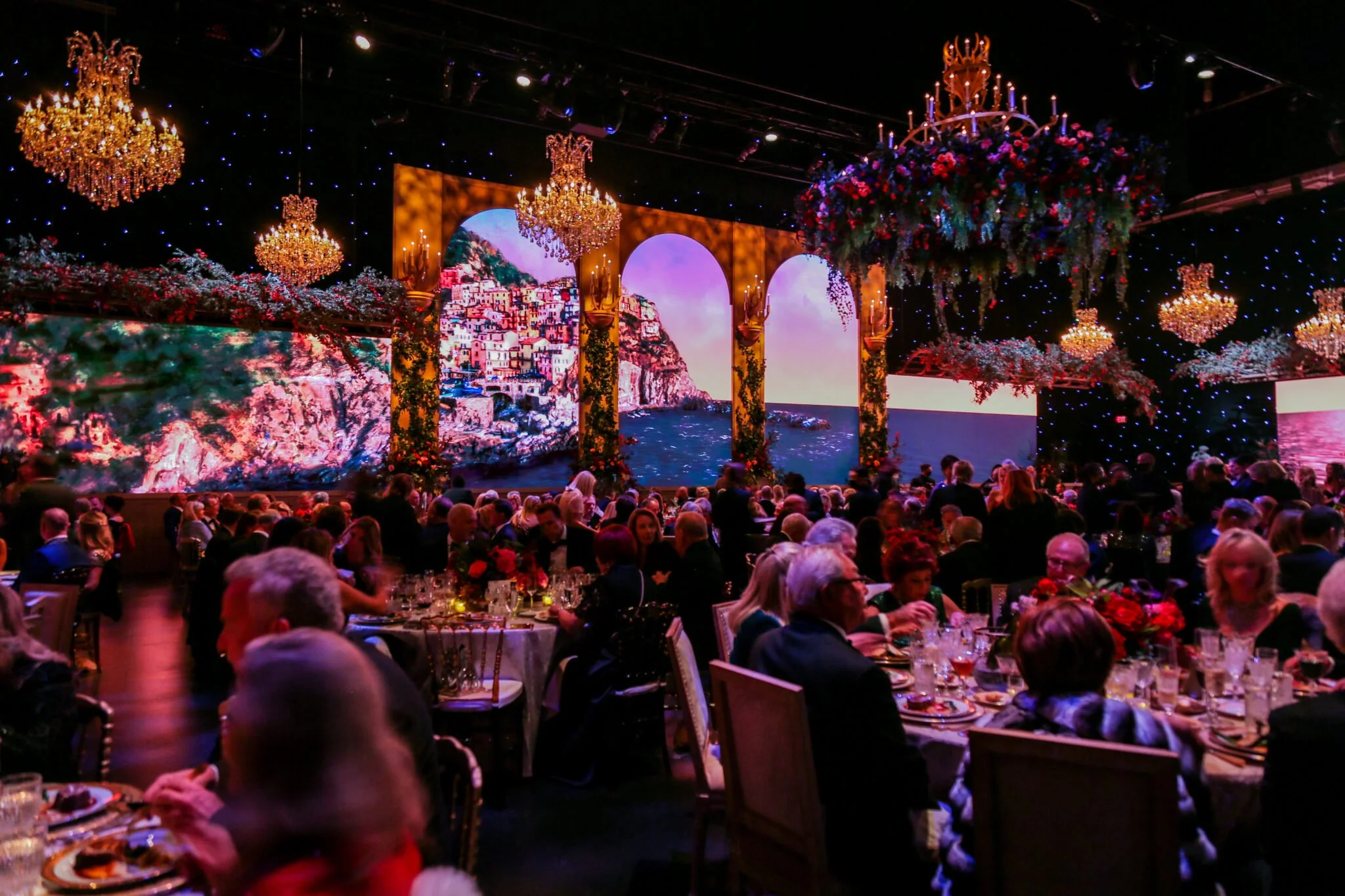 An indoor event with a large audience seated at tables set for dining. The background features a large digital screen displaying a picturesque coastal village scene with colorful buildings along a cliffside by the water. The ceiling is decorated with multiple chandeliers and large floral arrangements, creating an elegant ambiance.