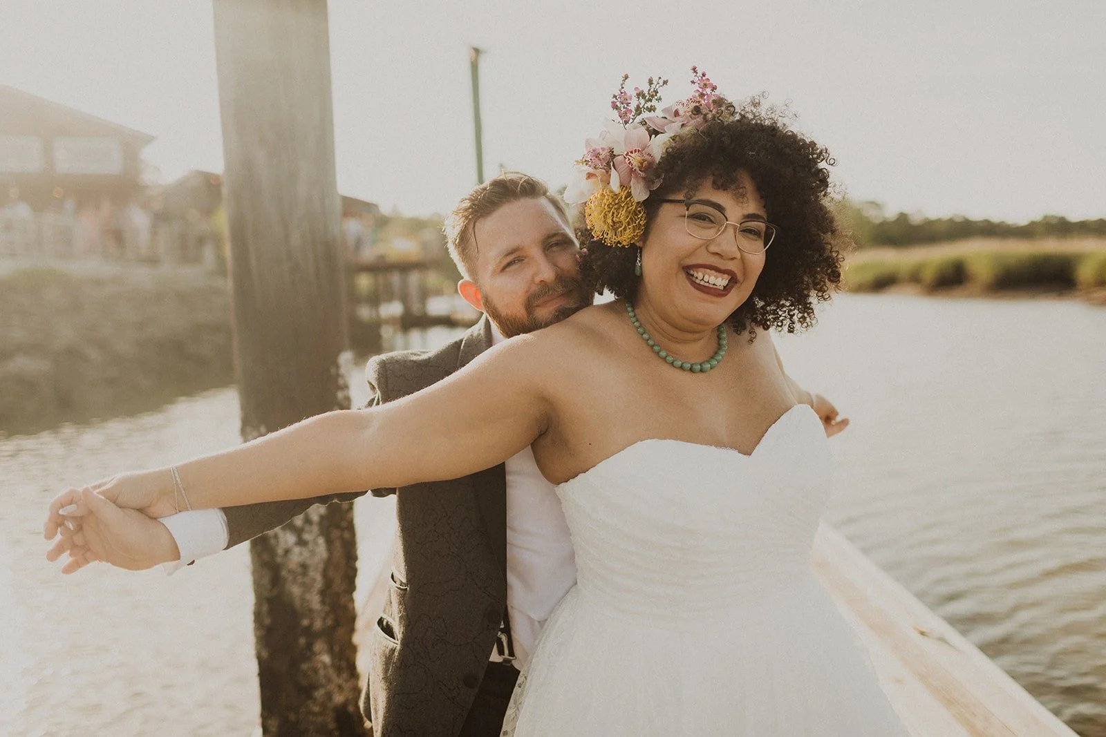 A joyful couple on their wedding day, with the groom embracing the bride from behind as she stands on a dock near the water, wearing a white strapless wedding dress and a floral headpiece.