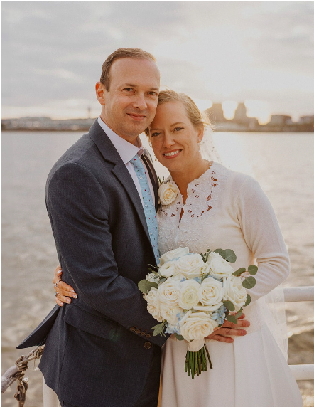A newlywed couple standing outdoors near a body of water, dressed in wedding attire. The bride is holding a bouquet of white roses, and the groom is wearing a suit with a blue tie. Both are smiling with the city skyline in the background during sunset.