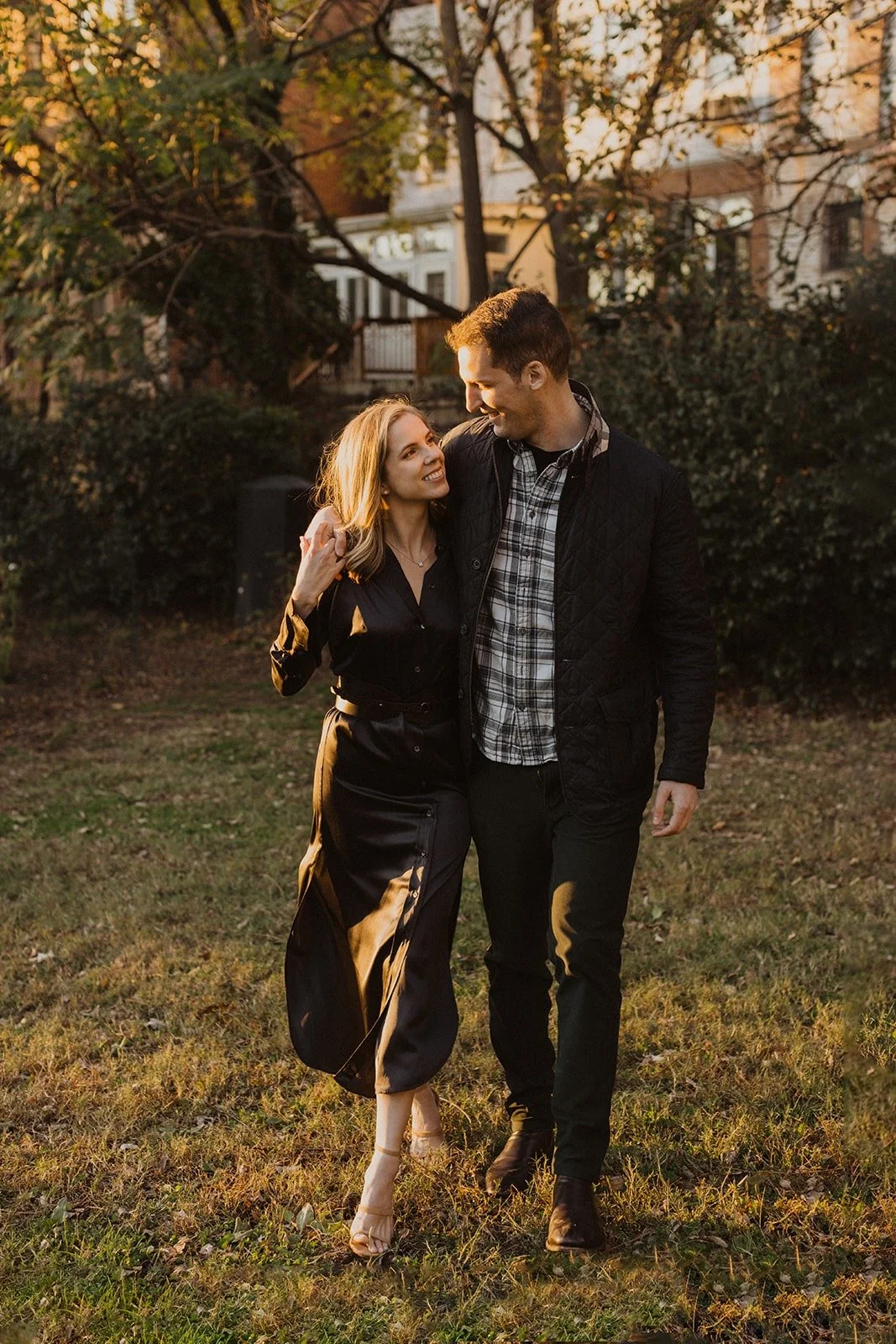 A young couple walking together outdoors in a park during the golden hour, smiling and looking at each other. The woman is wearing a black dress and beige heels, and the man is in a black jacket, plaid shirt, and black pants. They are surrounded by trees with some orange and yellow leaves.