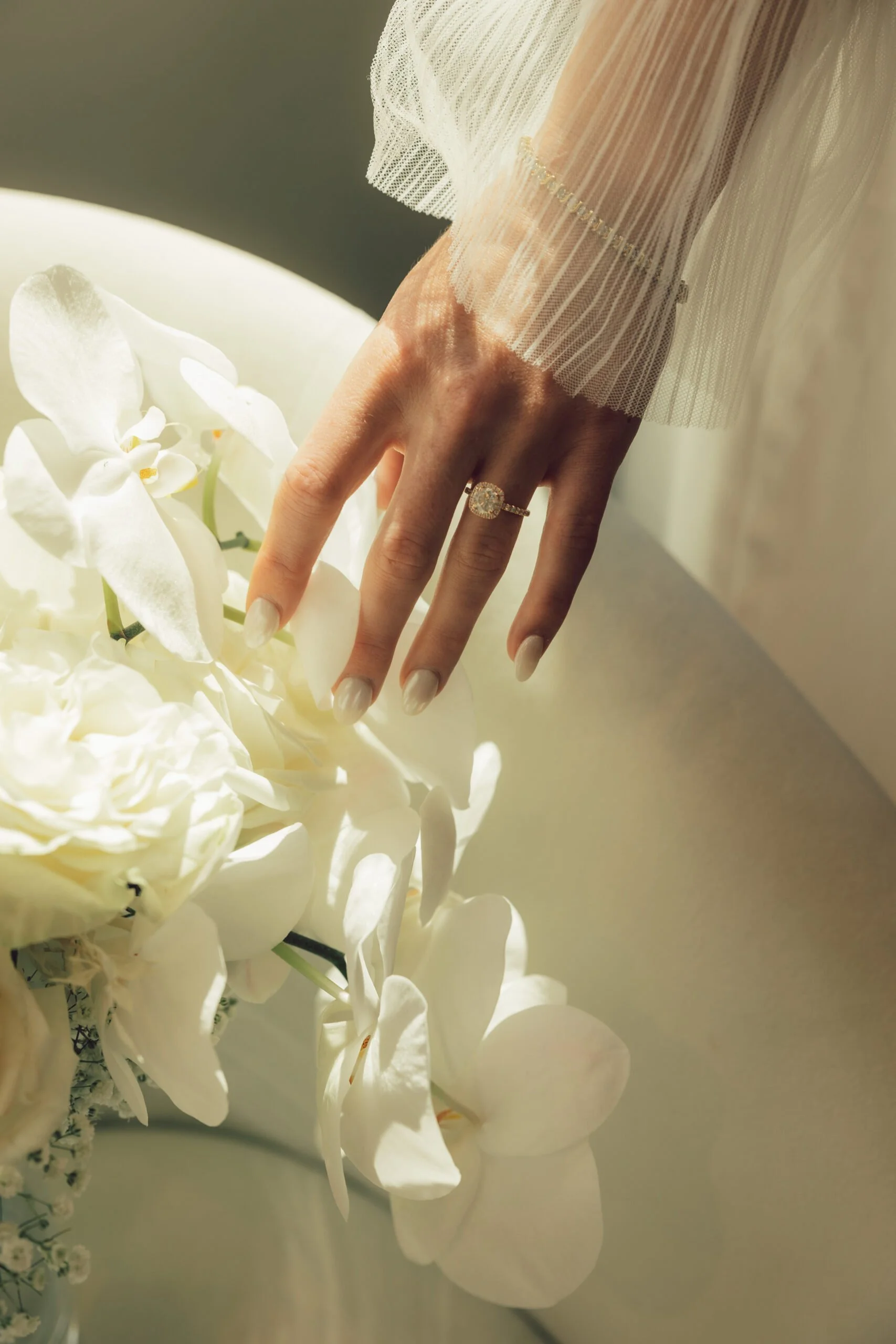 A close-up of a woman's hand with an engagement ring, resting on white orchids, with sheer fabric and lace details visible.