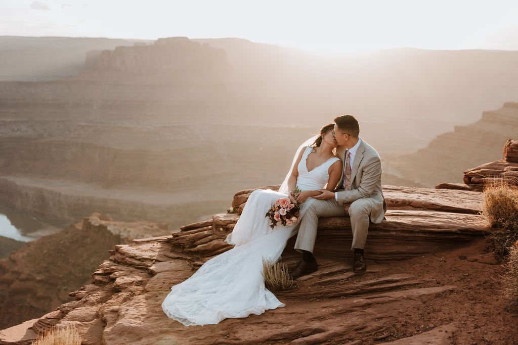 A bride and groom sharing a romantic moment on the edge of a cliff in a desert landscape during sunset.