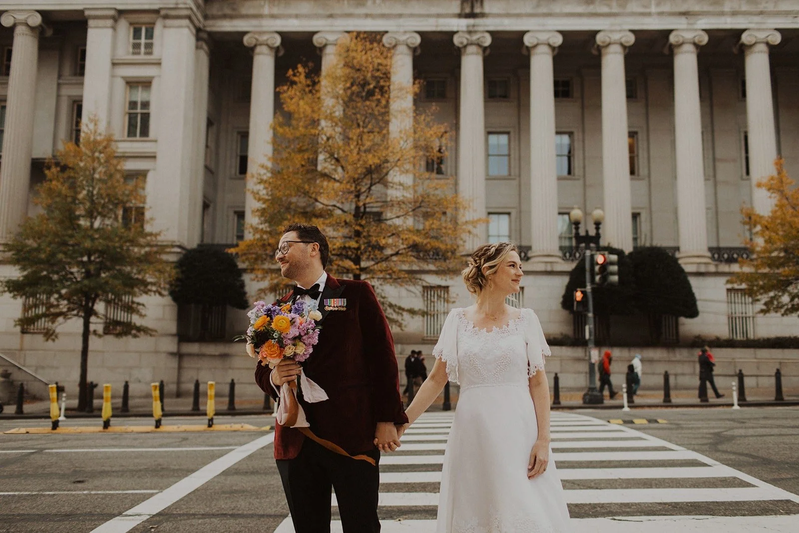 A bride and groom holding hands and standing at a crosswalk in front of a large neoclassical building with tall columns and autumn trees in New York City.
