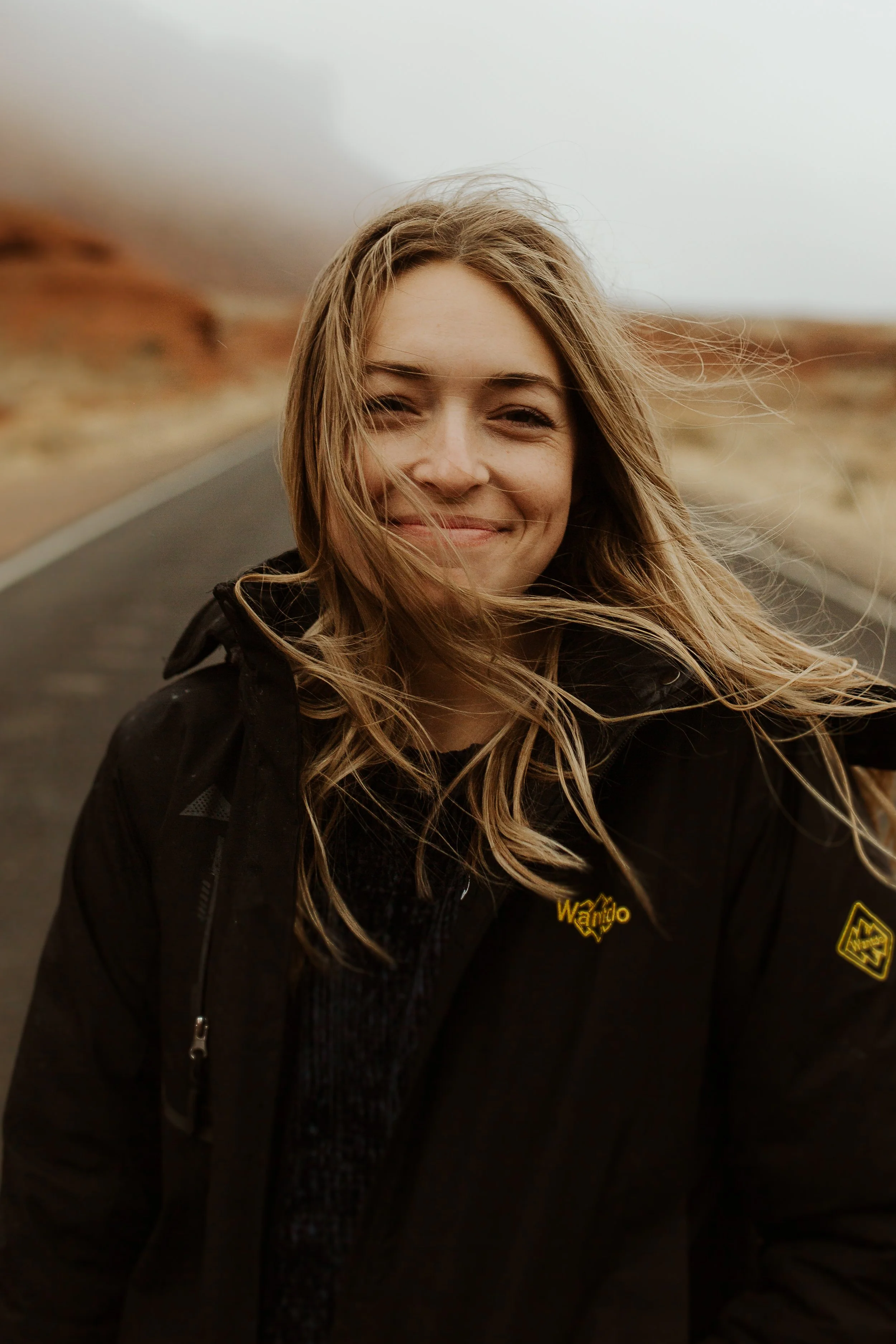 A smiling woman with long, windblown blonde hair standing on a rural road with a reddish-brown landscape and gray sky in the background.