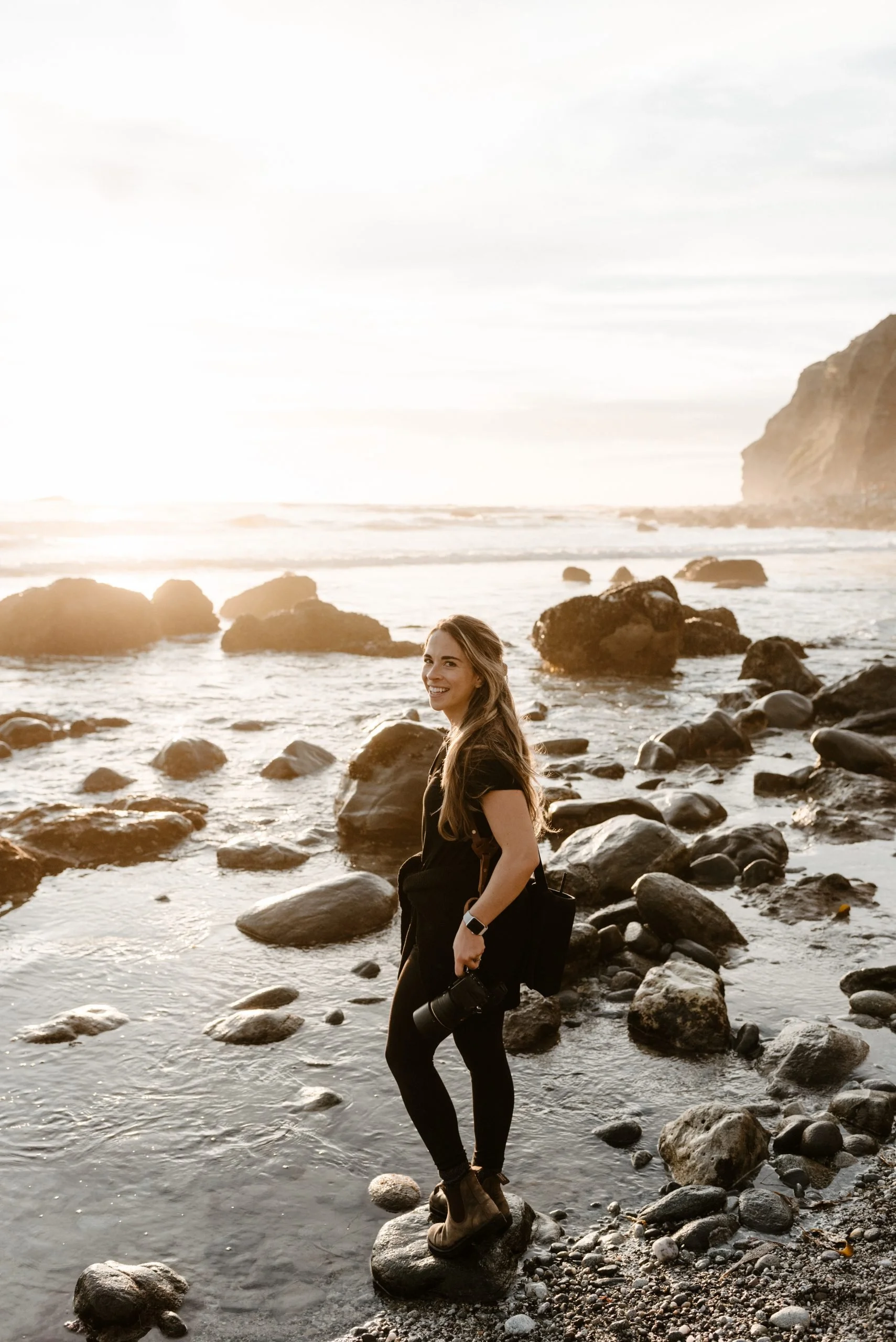A woman with long hair, dressed in black and wearing brown boots, is standing on rocks along a beach during sunset, holding a camera and smiling at the camera.
