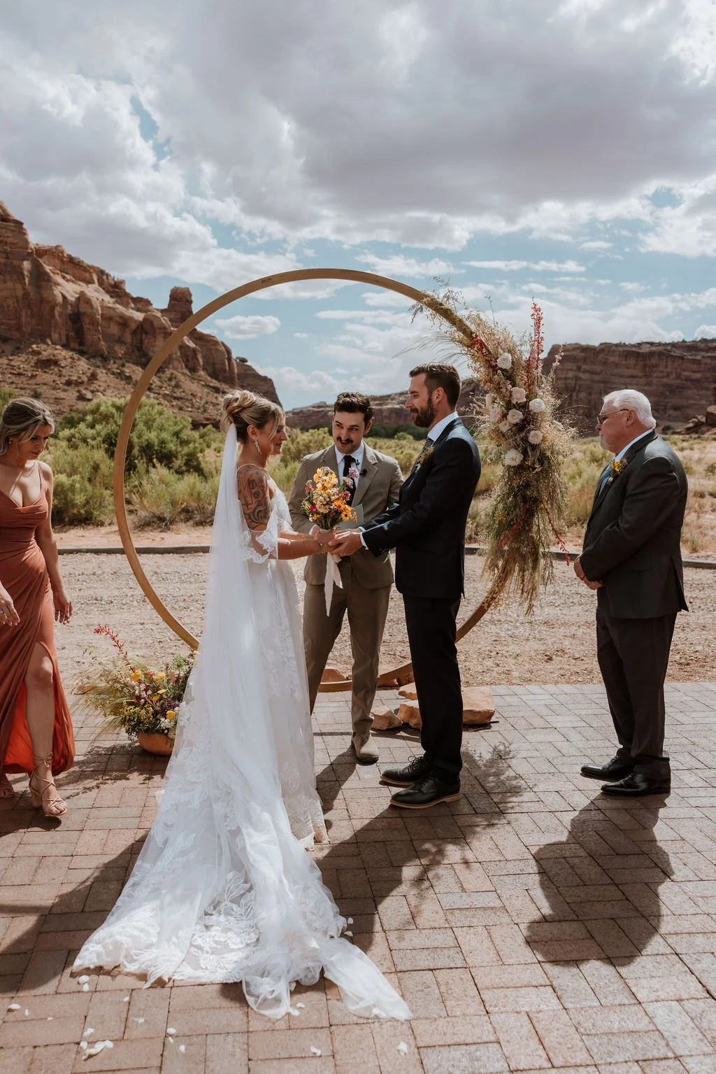 A wedding ceremony taking place outdoors in a desert setting with red rock formations in the background. The bride and groom are holding hands, exchanging vows, and smiling at each other, standing in front of a decorative circular arch adorned with flowers and greenery. An officiant stands between them, with a bridesmaid and a groomsman to the side.