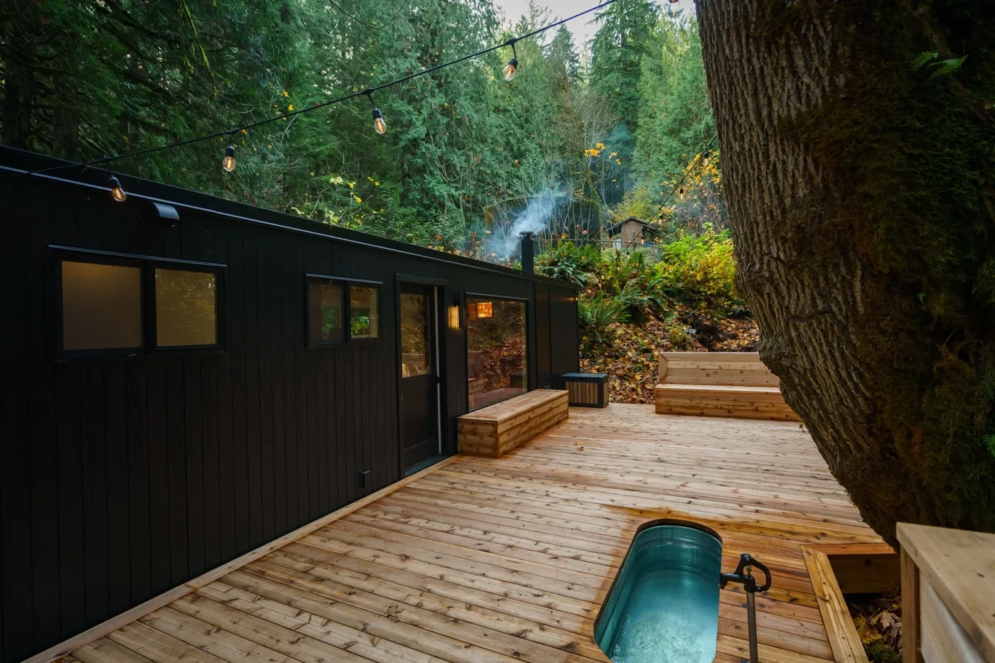 A modern black cabin with wood decking nestled in a lush forest with string lights hanging overhead.