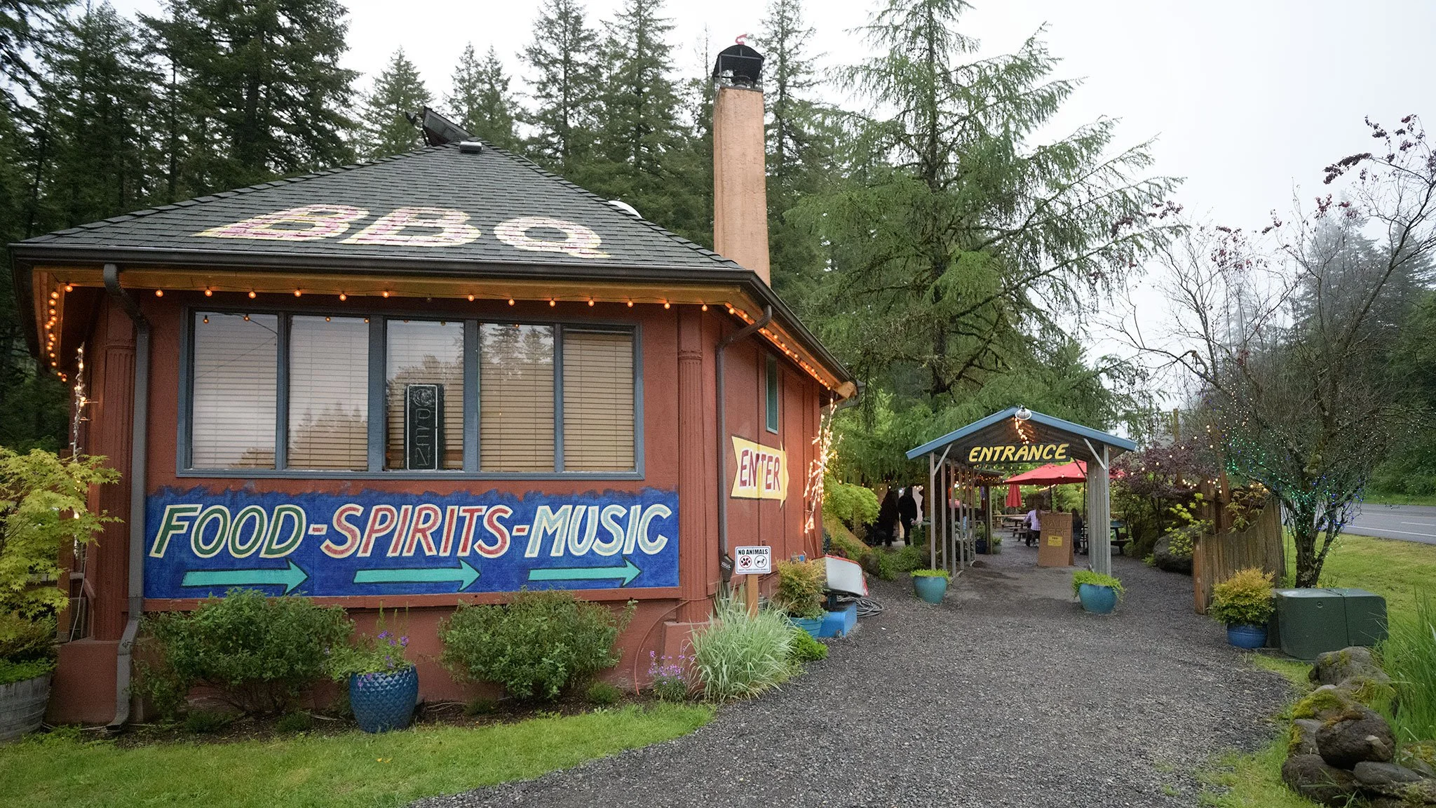 A rustic cafe or restaurant with a sign offering food, spirits, and music, decorated with outdoor string lights, surrounded by greenery, trees, and potted plants, with an entrance marked by a blue awning and outdoor seating area.