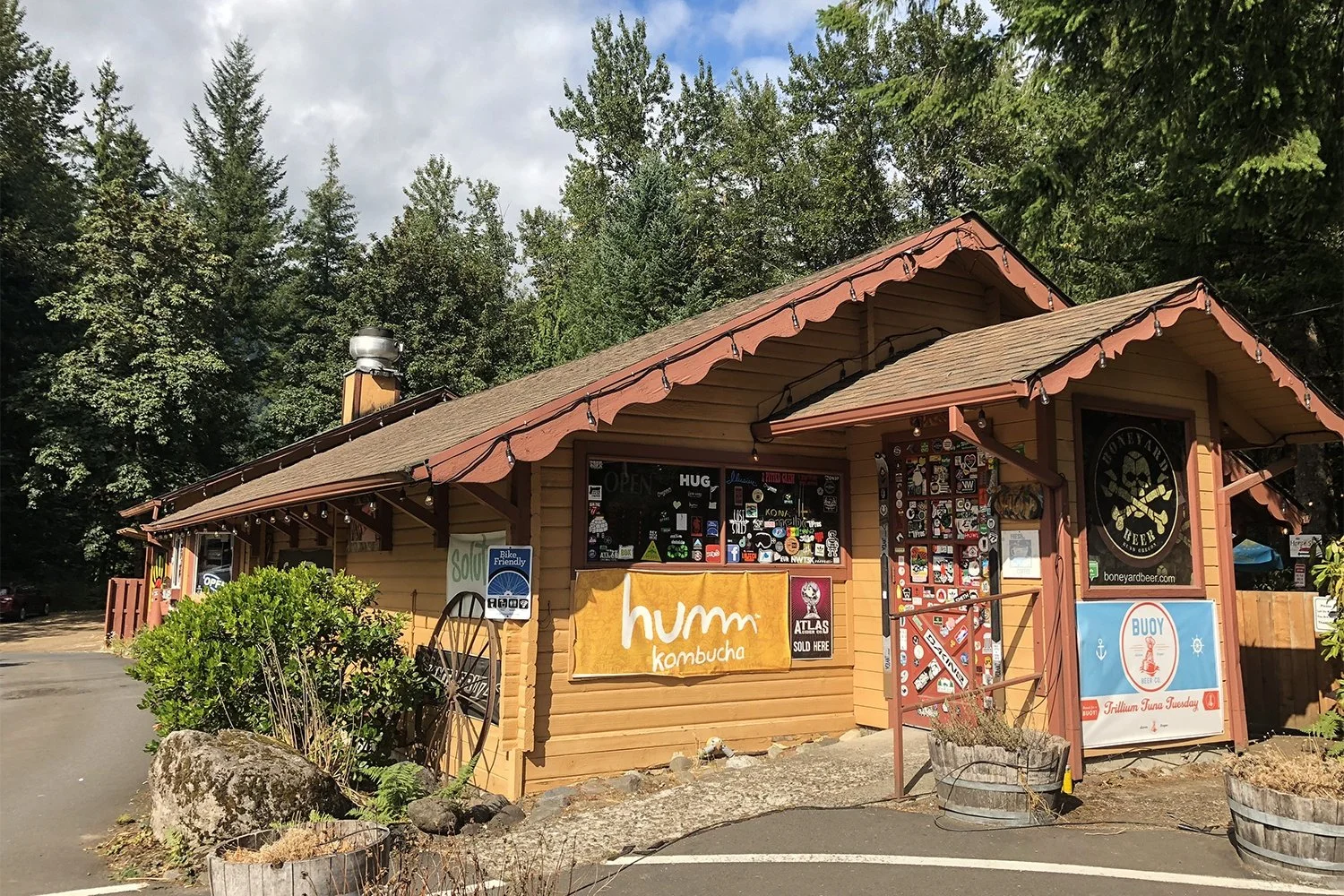 A small wooden building with a brown roof, decorated with string lights, surrounded by Trees; signs advertising kombucha, beer, and other products are on the building's exterior.