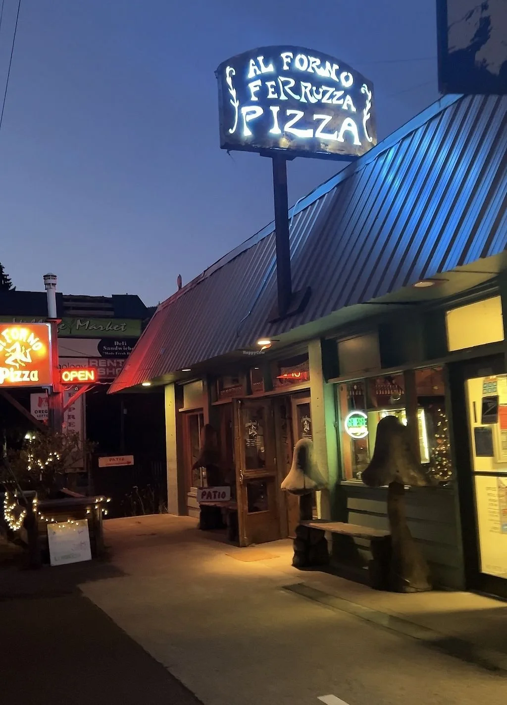 Nighttime outside view of a pizza restaurant with a sign that says "Sal Fermo Ferruzza Pizza" illuminated overhead. The restaurant has a green exterior with outdoor lighting, and signs indicating it is open. There are decorative mushroom sculptures near the entrance and other nearby signs and lights.