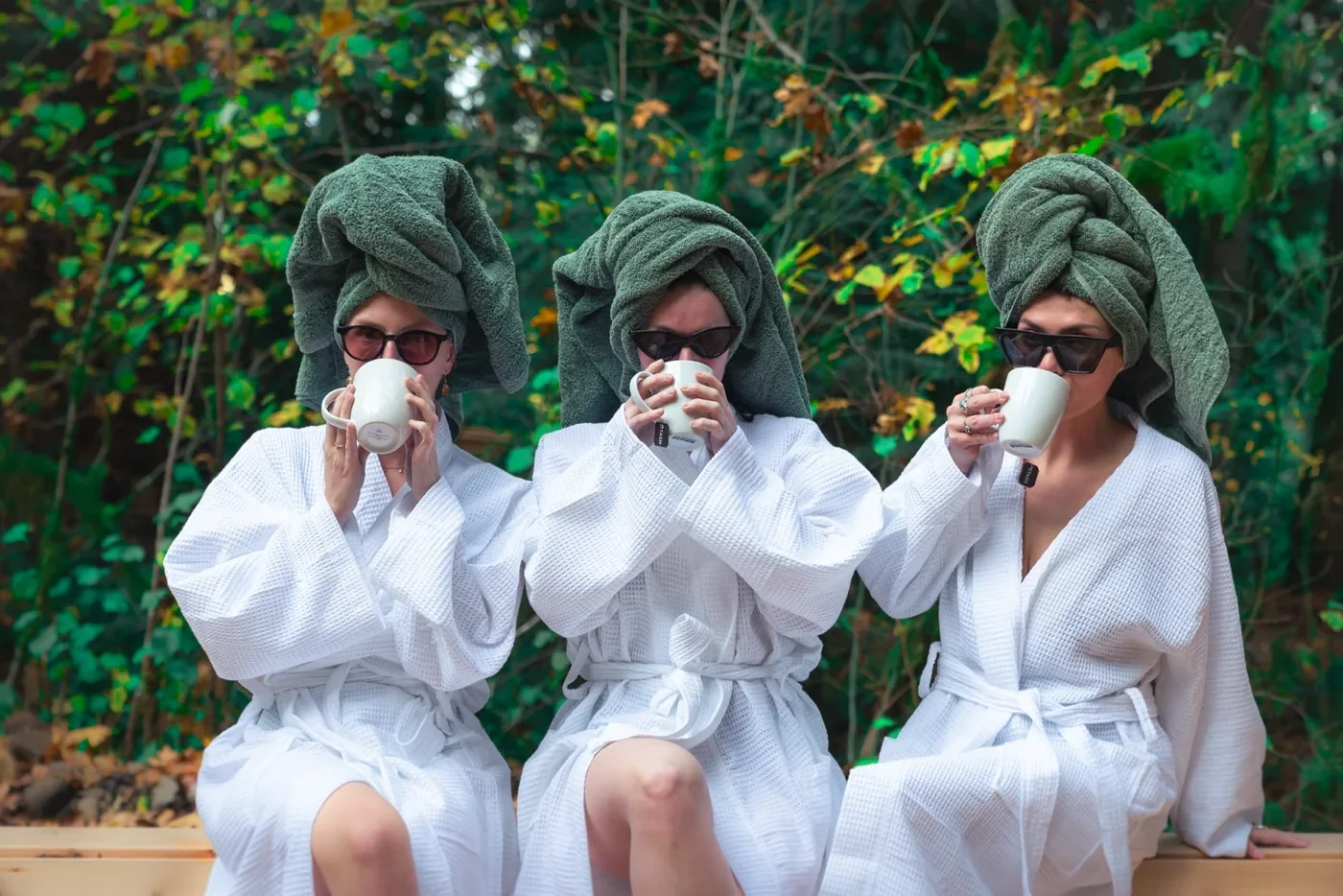 Three women sitting outdoors in robes with towels on their heads, drinking from white mugs, in front of green foliage.