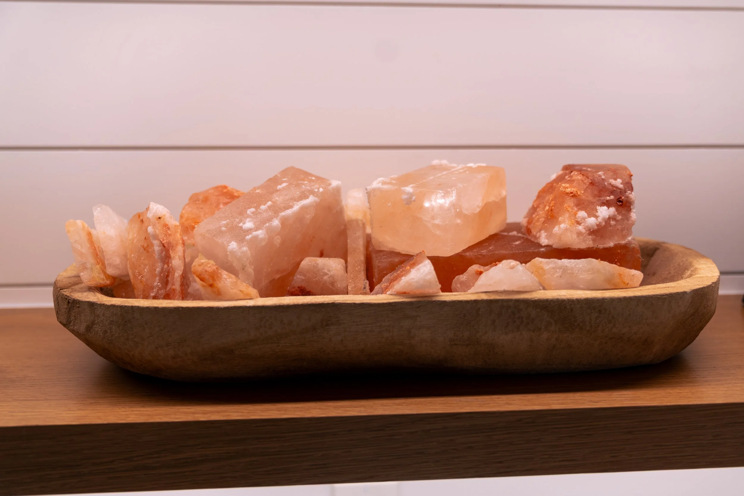 A wooden bowl filled with Himalayan salt stones on a wooden surface against a white paneled wall.
