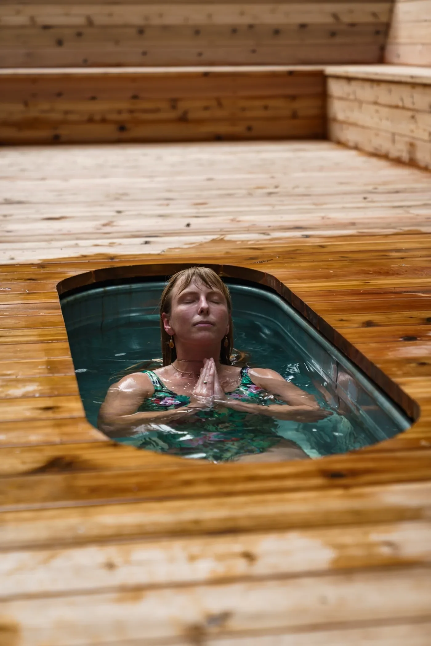 A woman meditating in a hot tub with her hands in prayer position, inside a wooden spa with wooden paneling.