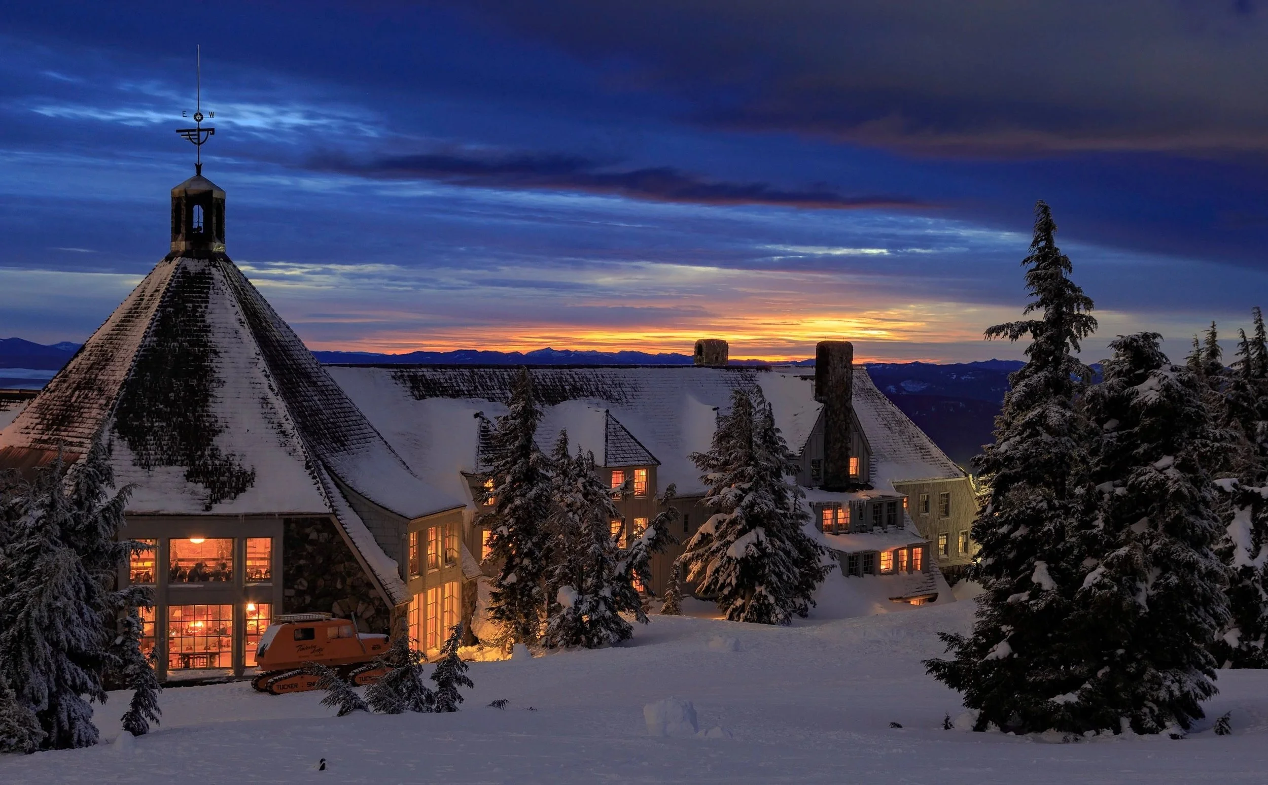 A snow-covered mountain lodge at dusk with lit windows, surrounded by snow-laden trees, under a colorful evening sky with clouds and a sunset in the background.