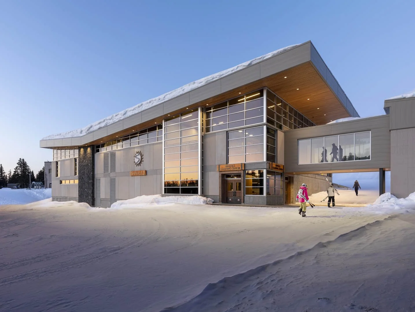 Modern building with large glass windows, snow on the roof and ground, and people skiing and walking nearby in a winter setting.