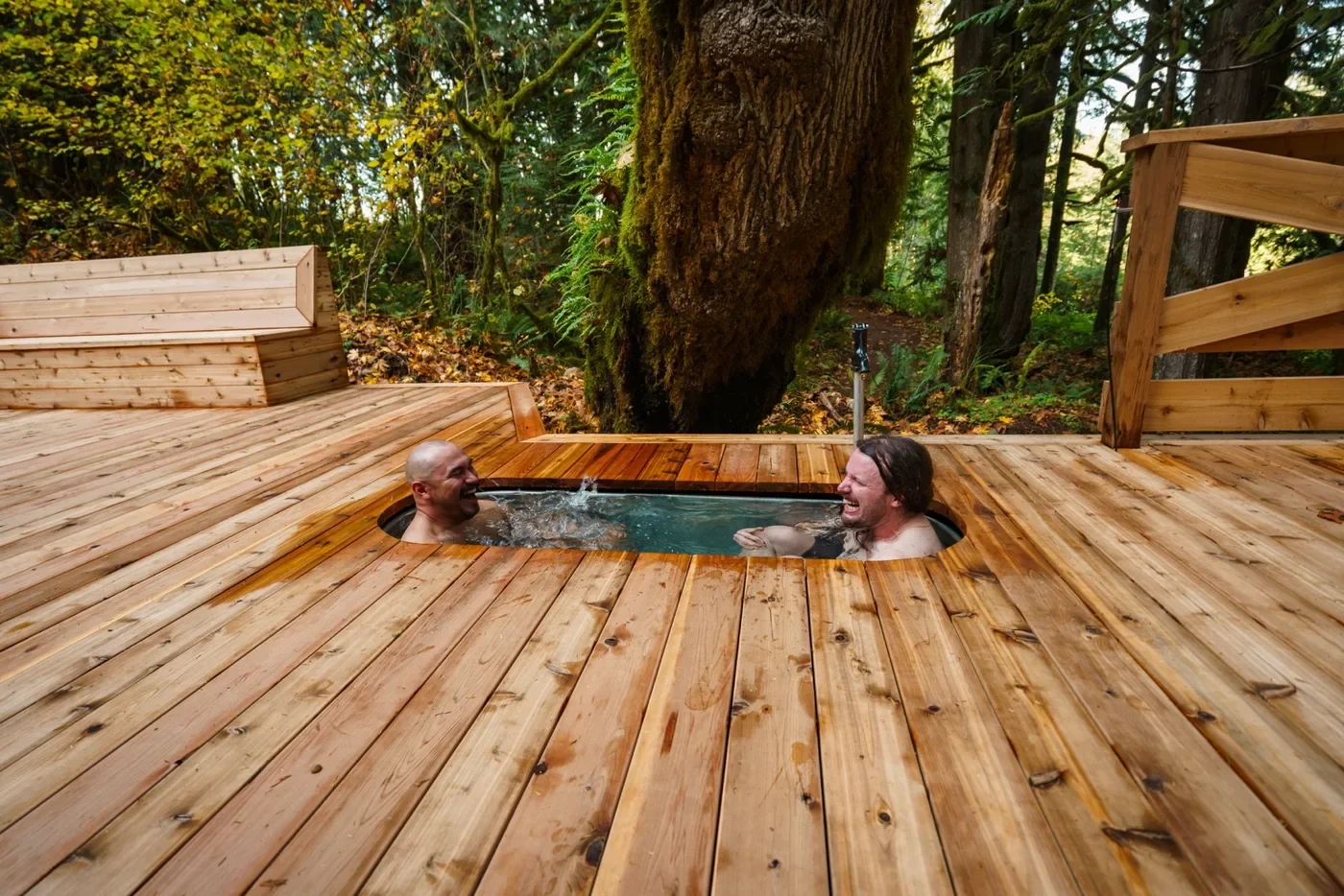 Two men laughing and relaxing in a small outdoor hot tub surrounded by a wooden deck in a forested area.