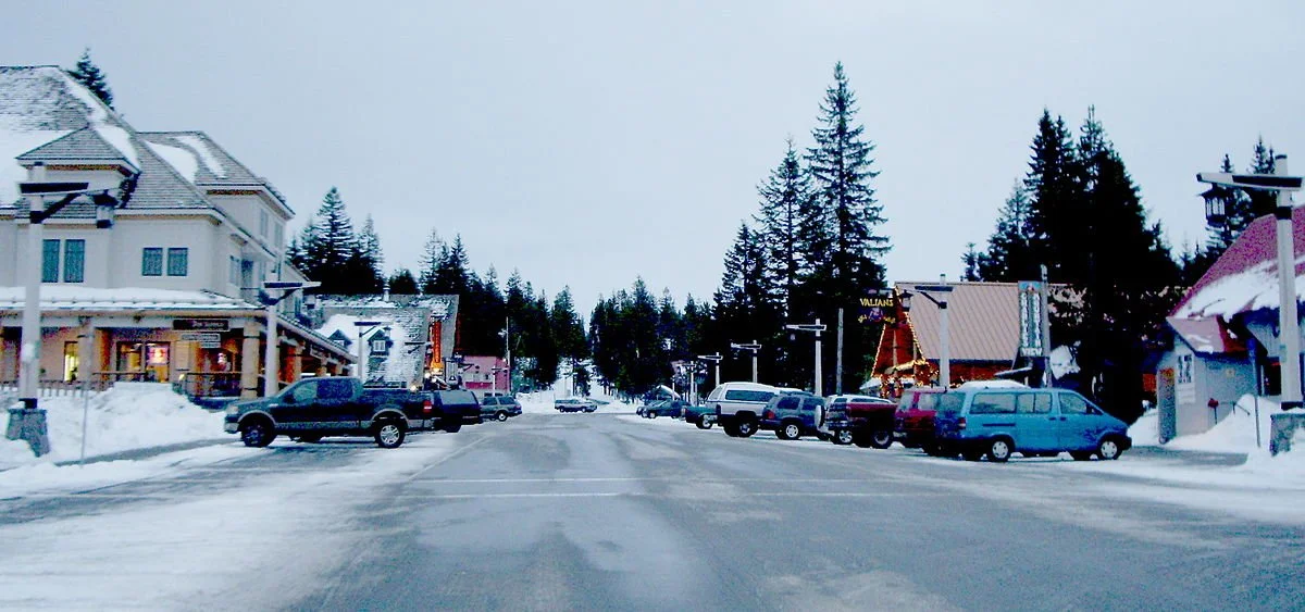A snowy parking lot in a small town with cars parked on both sides, surrounded by buildings and evergreen trees, in winter.