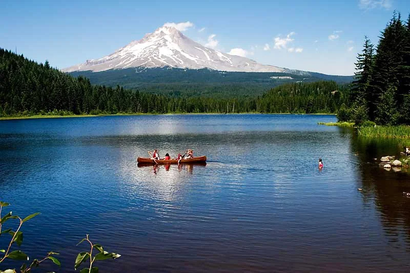 A lake with a canoe carrying four people, surrounded by dense green trees with a snow-capped mountain in the background under a blue sky.