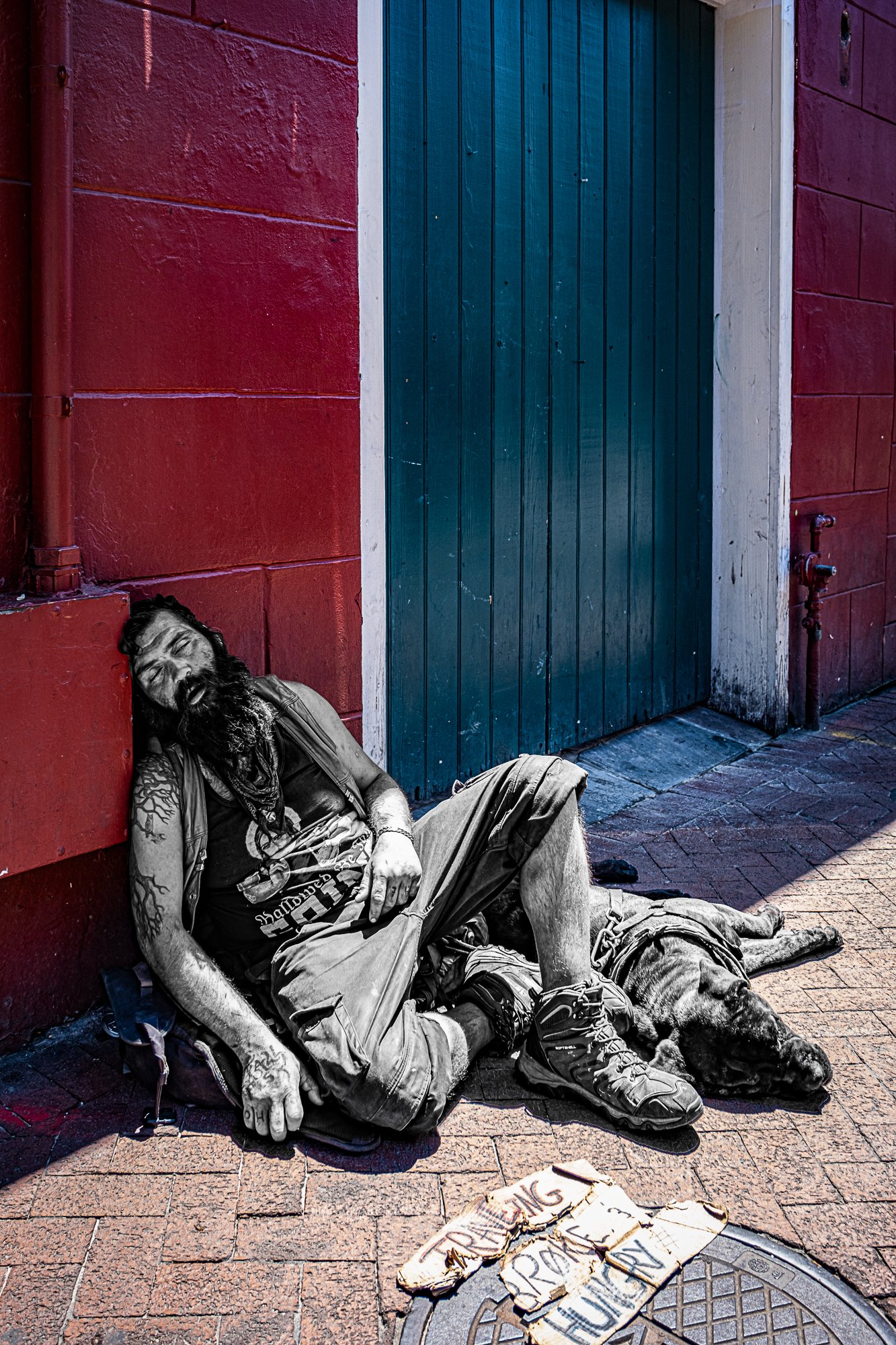 On the edge of a red wall, a man sleeps with his head resting on a box, propped up against a building. His dog is curled beside him, peaceful and still. A cardboard sign reads: traveling, broke, and hungry. People pass without stopping. Some glance.