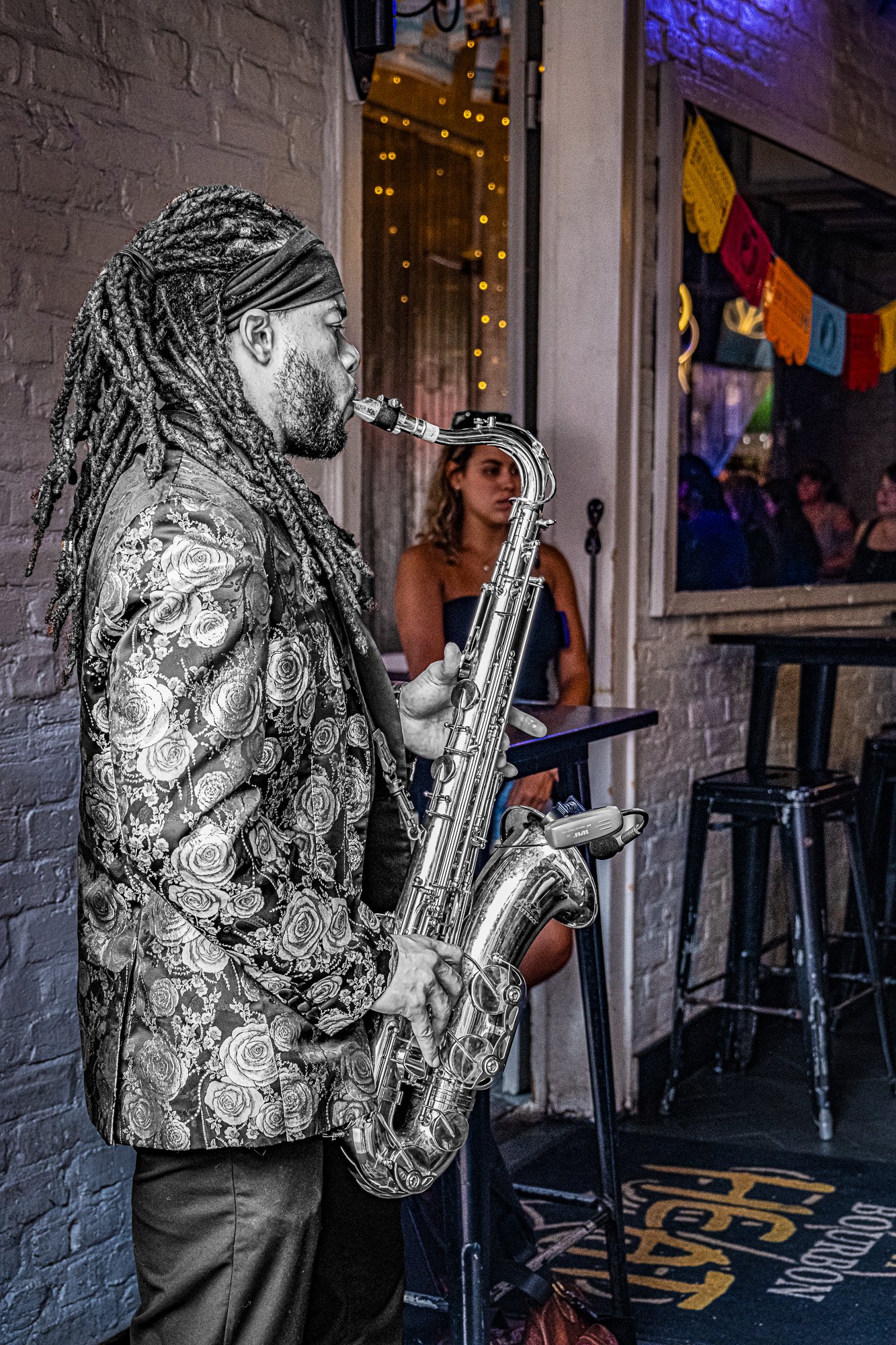 Just inside the entrance of a bar, a saxophonist stands playing, hoping his sound will spill into the street and draw people in. He plays with purpose—every note a pitch for attention, every glance toward the sidewalk a quiet ask. He’s performing for