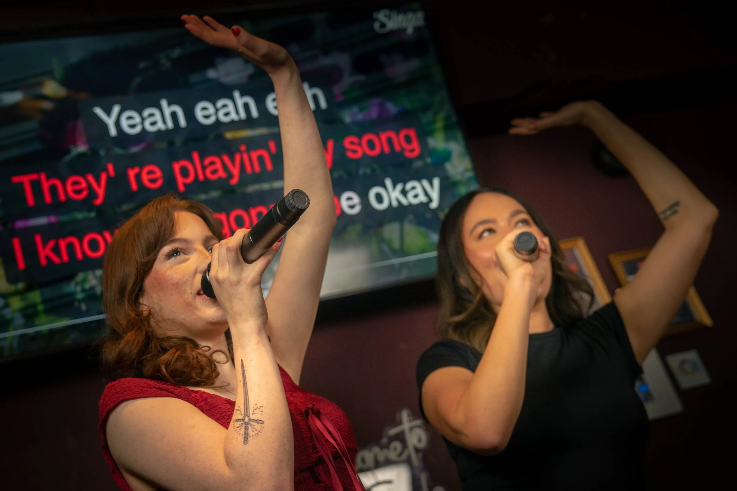 Two women singing into microphones on stage with karaoke lyrics displayed on a screen behind them. One woman in a red dress has her arm raised and the other in a black shirt has her hand near her head.