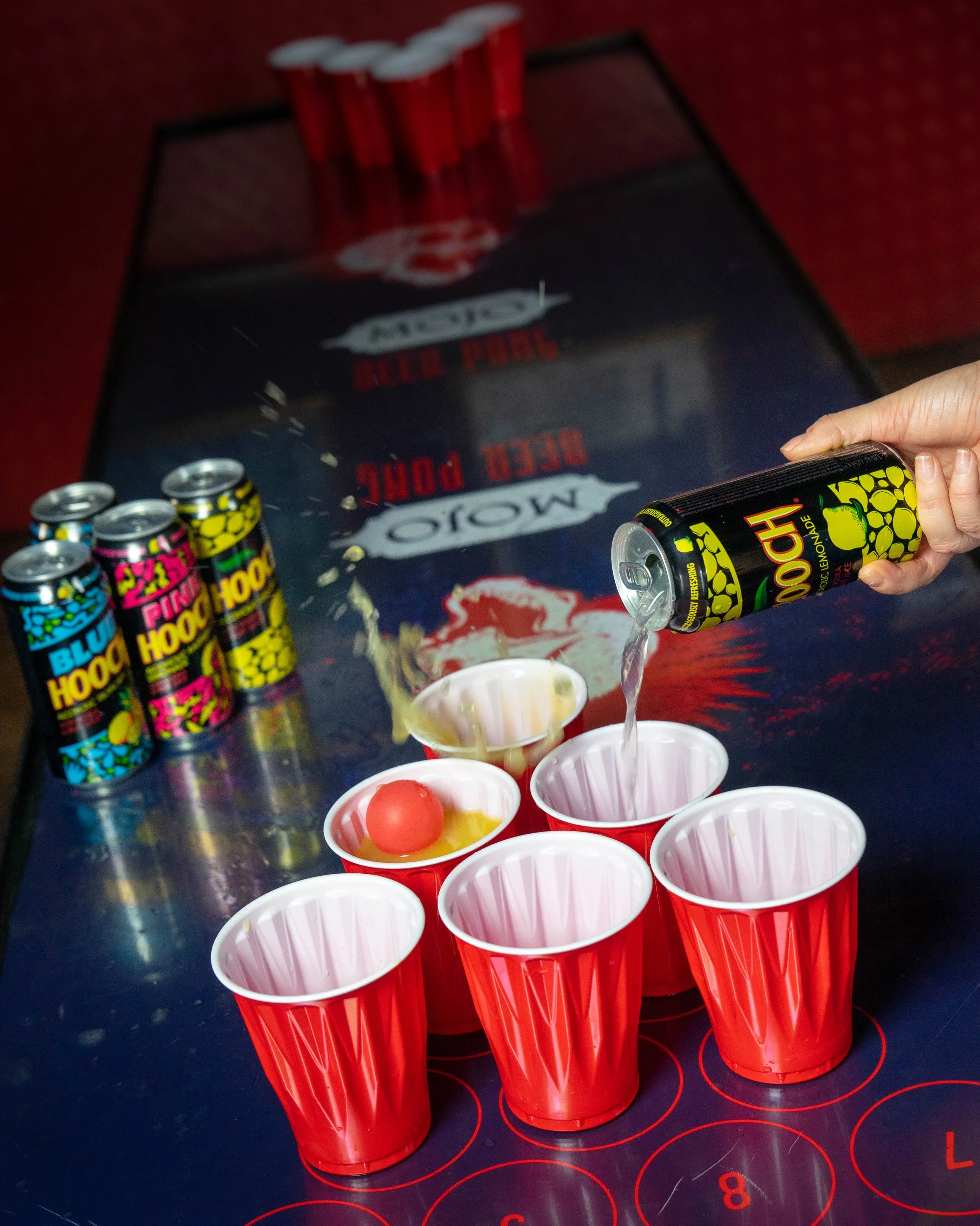A person pouring a pink soda into red plastic cups with one egg in one of the cups. Several cans of colorful soda are on the table, and a table with writing and a logo in the background.