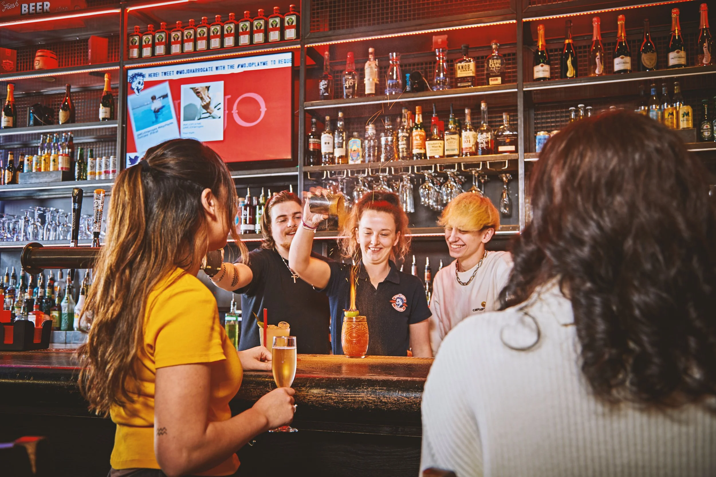 A bar scene with four people socializing and a bartender preparing drinks. The bartender is pouring a drink into a glass, and another person is holding a champagne flute. The bar is stocked with various bottles of alcohol and glasses, and a digital screen is visible in the background.