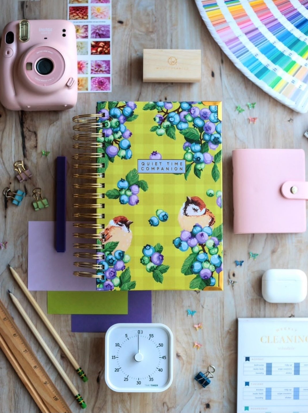 Flat lay of a wooden desk with a colorful planner titled "Quiet Time Companion" featuring birds and berries, a purple pen, a blue pen, a ruler, a sticky note pad, pastel binder clips, a pink instant camera, a color wheel, a small wooden block, a pink