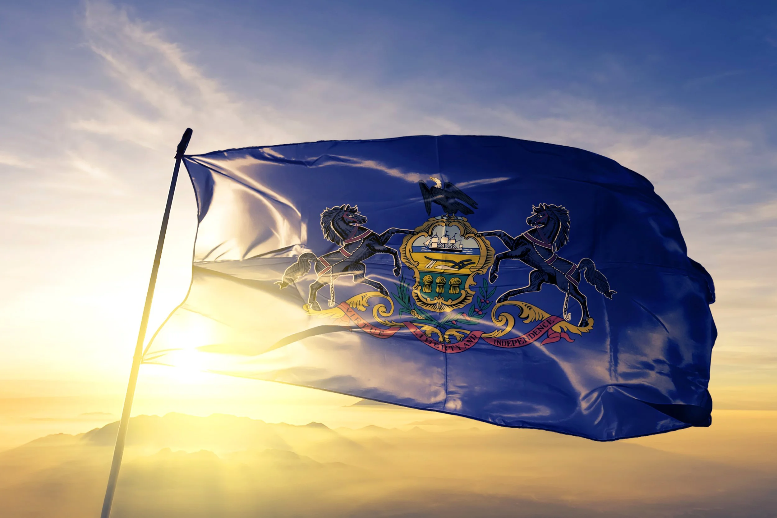 The flag of Pennsylvania waving in the wind during sunset with mountains and a cloudy sky in the background.