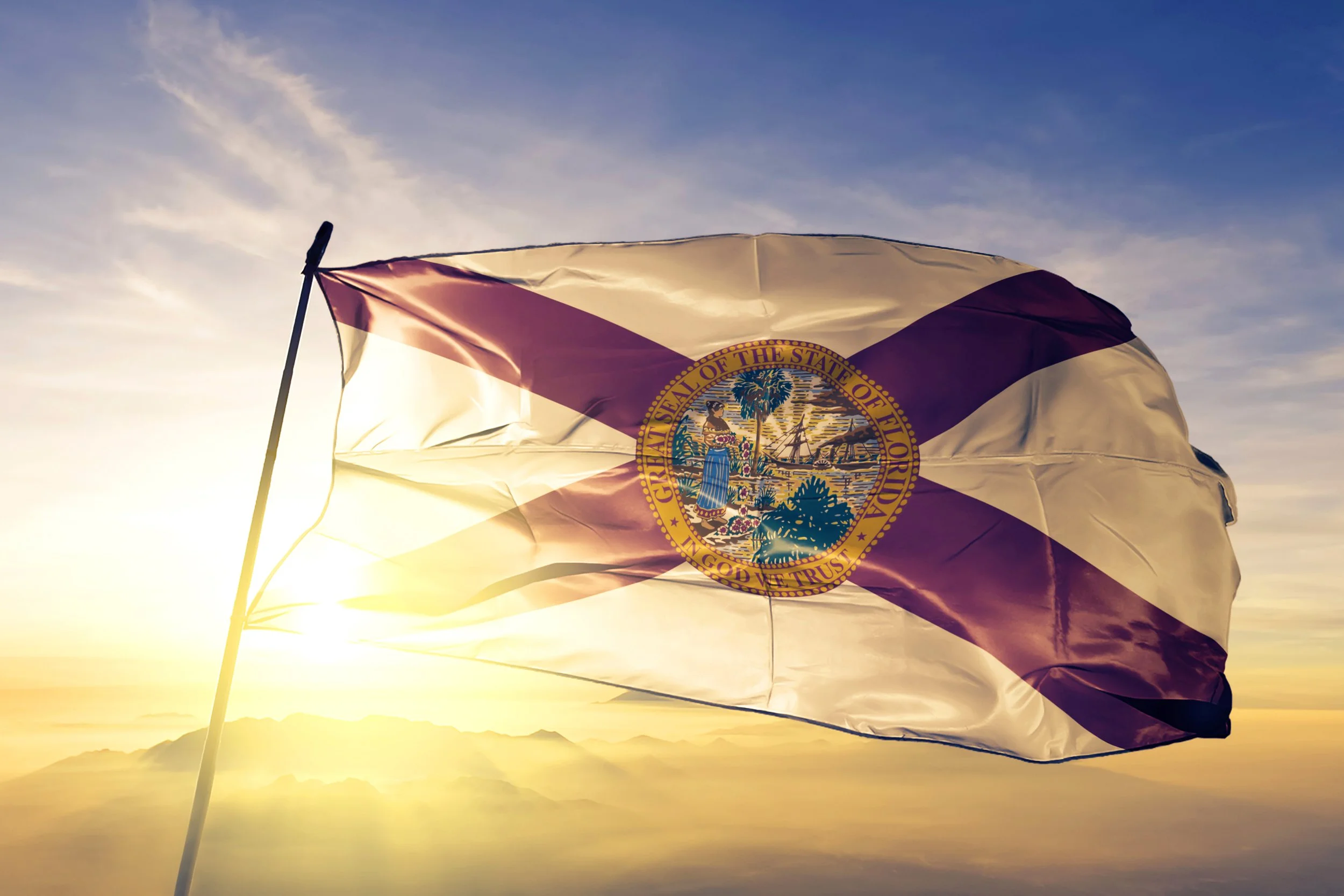 The Florida state flag fluttering against a sunset sky with mountains and clouds in the background.