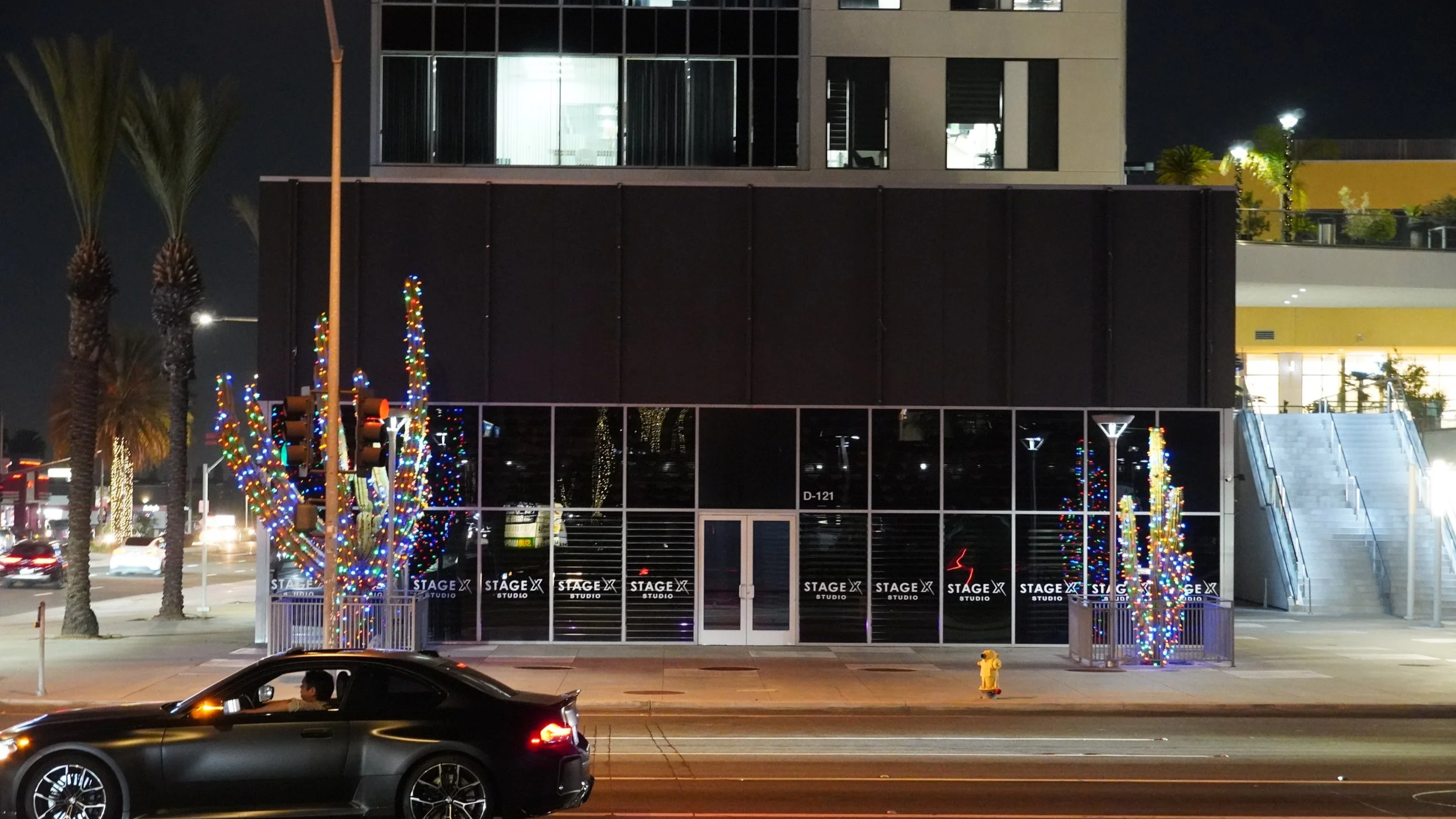 Nighttime street scene with a modern black building, colorful holiday lights on trees, a black car driving by, and a yellow fire hydrant on the sidewalk.