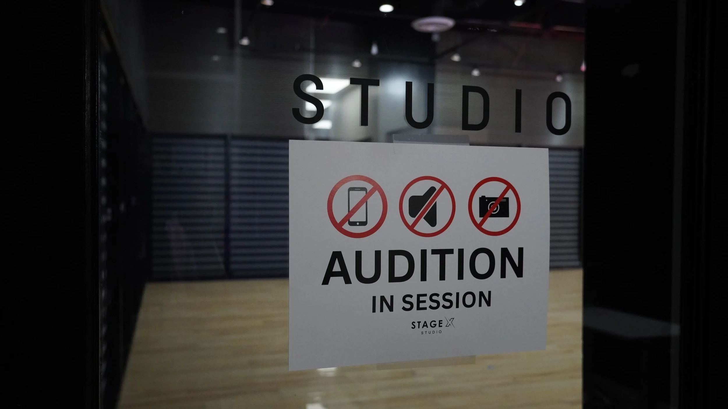 Sign on glass door of a dance studio indicating that the audition is in session and listing prohibited items such as cell phones, folders, and cameras.
