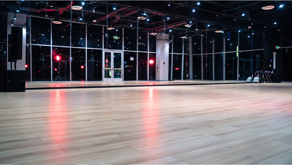 Empty dance studio with a large mirrored wall, wooden floor, and colorful string lights overhead.