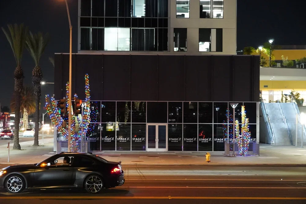 Night view of a modern building with entrance doors, decorated trees with colorful lights, a black car on the street, and palm trees in the background.