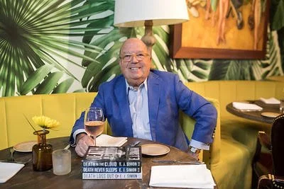 Author E.J. Simon with a glass of wine and three books in front of palm fronds