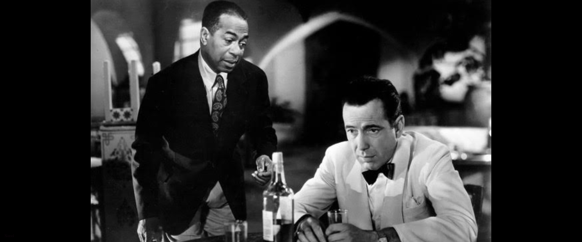 A man sitting at a table drinking liquor with a man standing next to him
