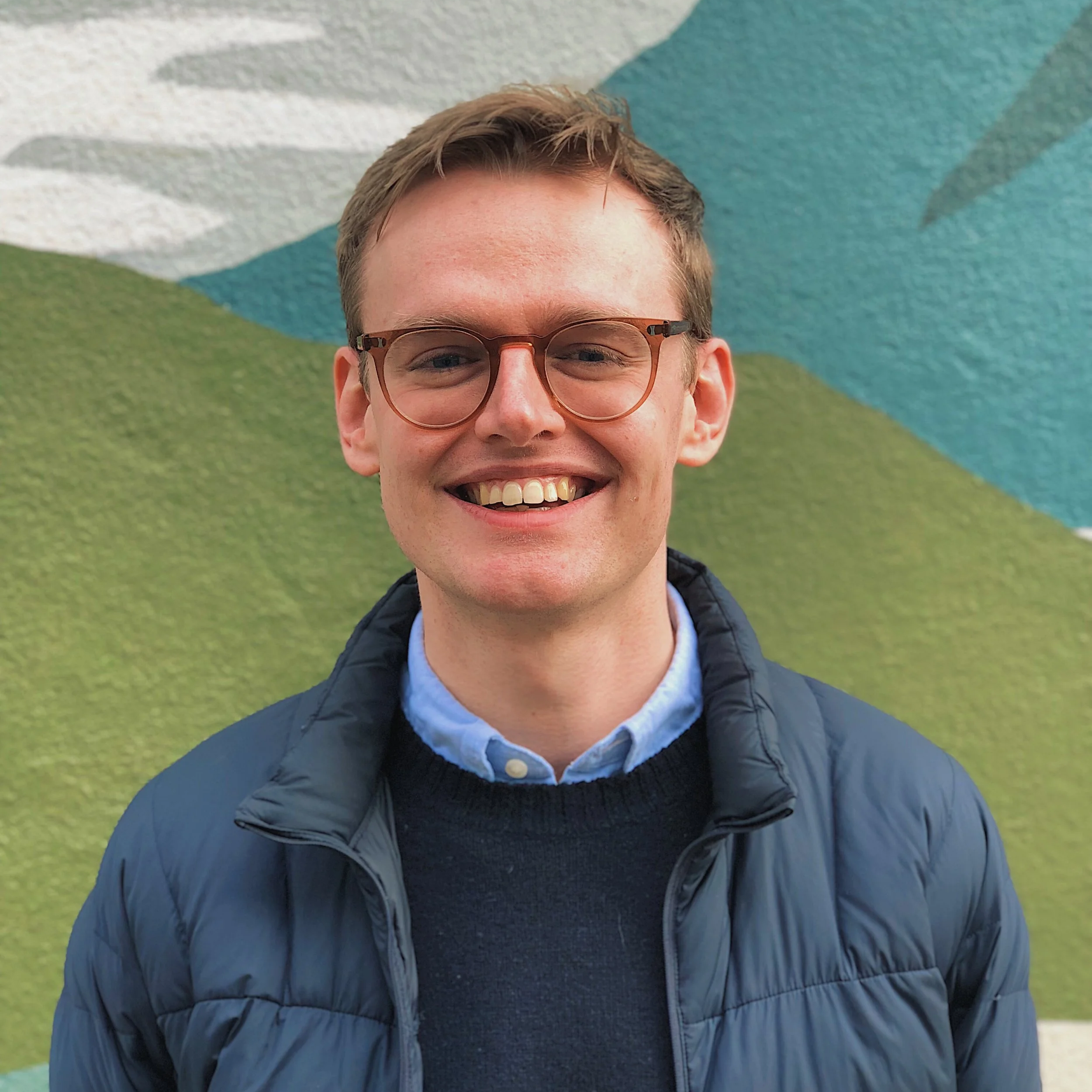 A young man with glasses smiling outdoors with a colorful mural in the background.