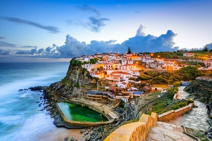 Coastal village on a cliff with buildings, a swimming pool, and ocean waves during sunset.