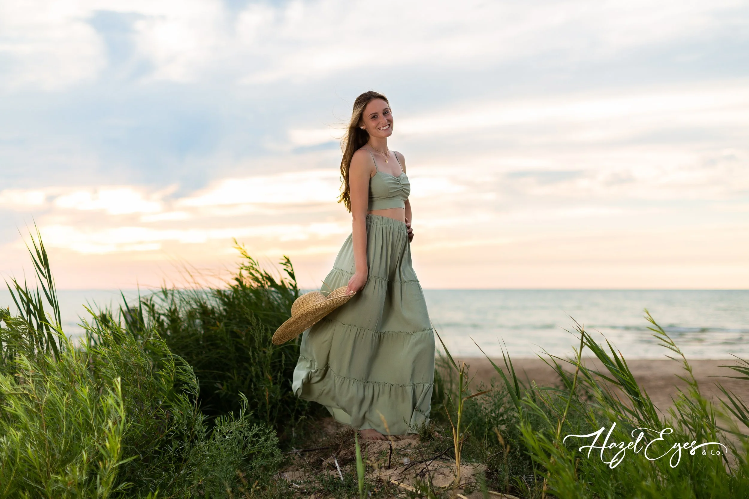 High school senior girl on the beach with hat in hand blowing in the wind.