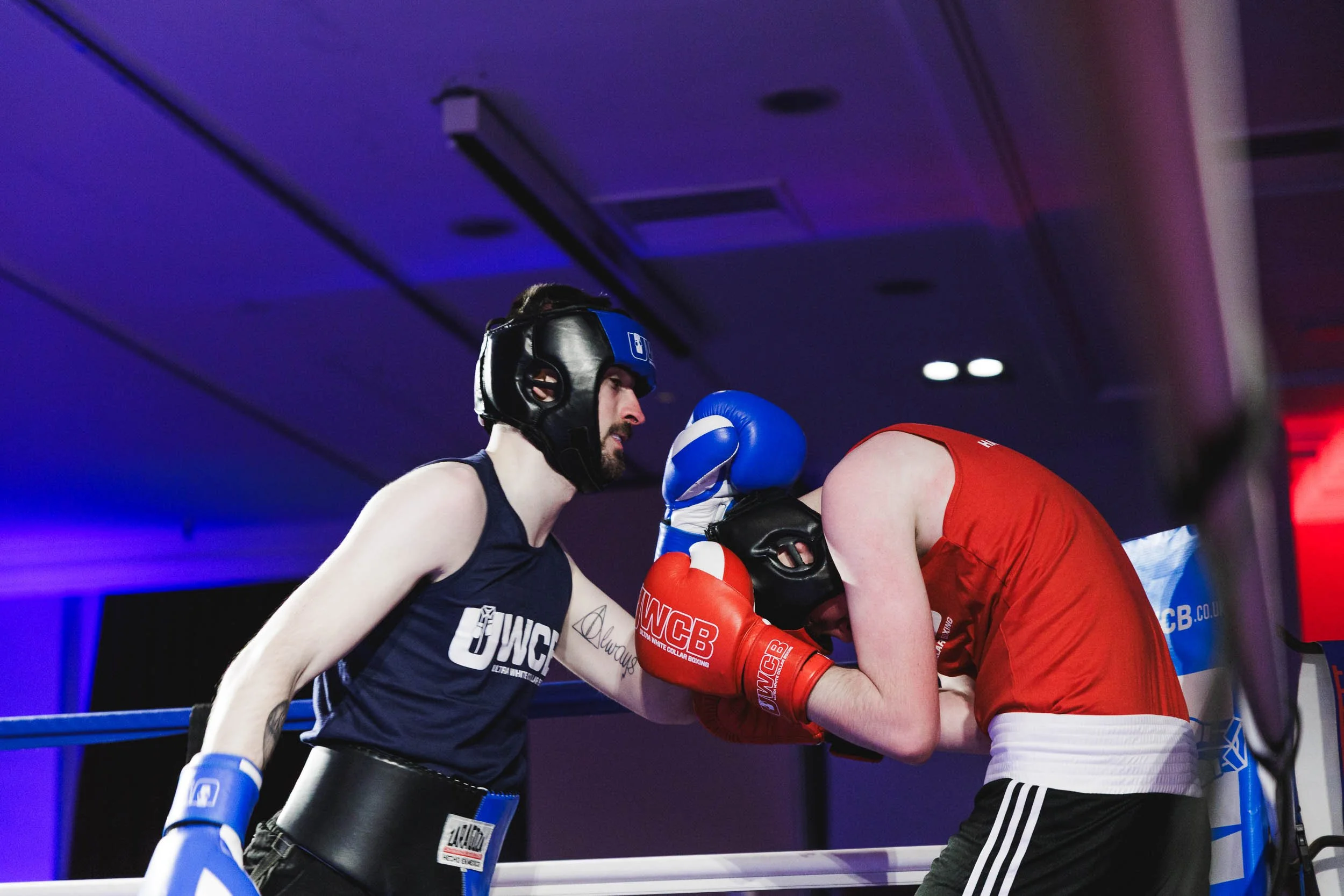 Two male amateur boxers in a boxing ring during a match, one wearing a blue helmet and gloves, the other wearing a red helmet and gloves, engaged in a fight with the red-gloved boxer leaning forward.