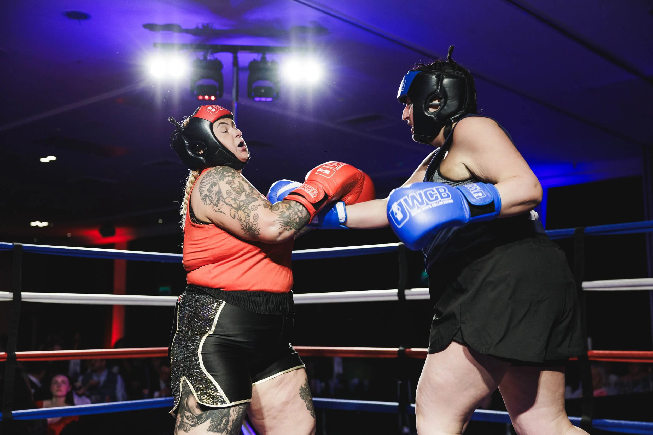 Two female boxers wearing protective headgear and gloves are fighting in a boxing ring. One is in red gloves and the other in blue gloves, and they are engaging in a close exchange of punches.