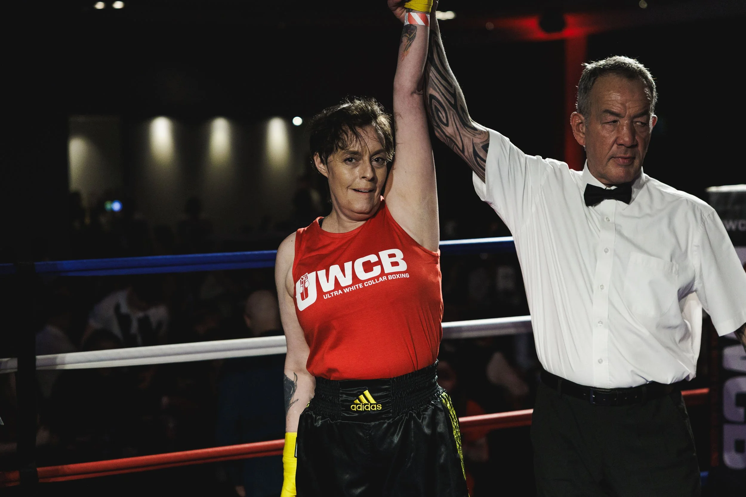 A female boxer in a red tank top with the text 'UWCB Ultra White Collar Boxing' is being declared the winner in a boxing match, with a referee raising her arm.