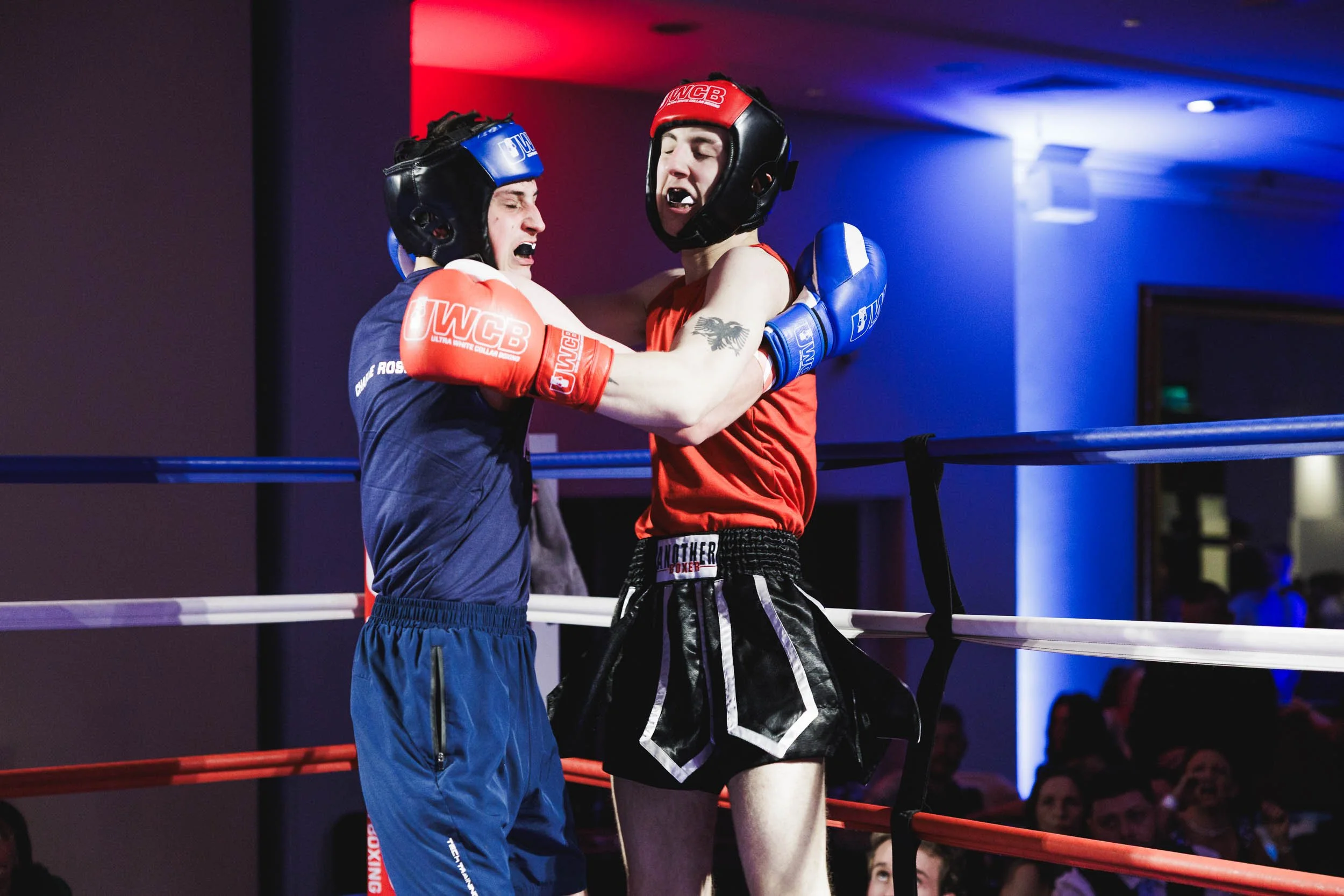 Two male amateur martial artists engaged in a boxing match inside a boxing ring, wearing protective headgear and gloves. One is in red gear, the other in blue, with a competitive environment and audience in the background.