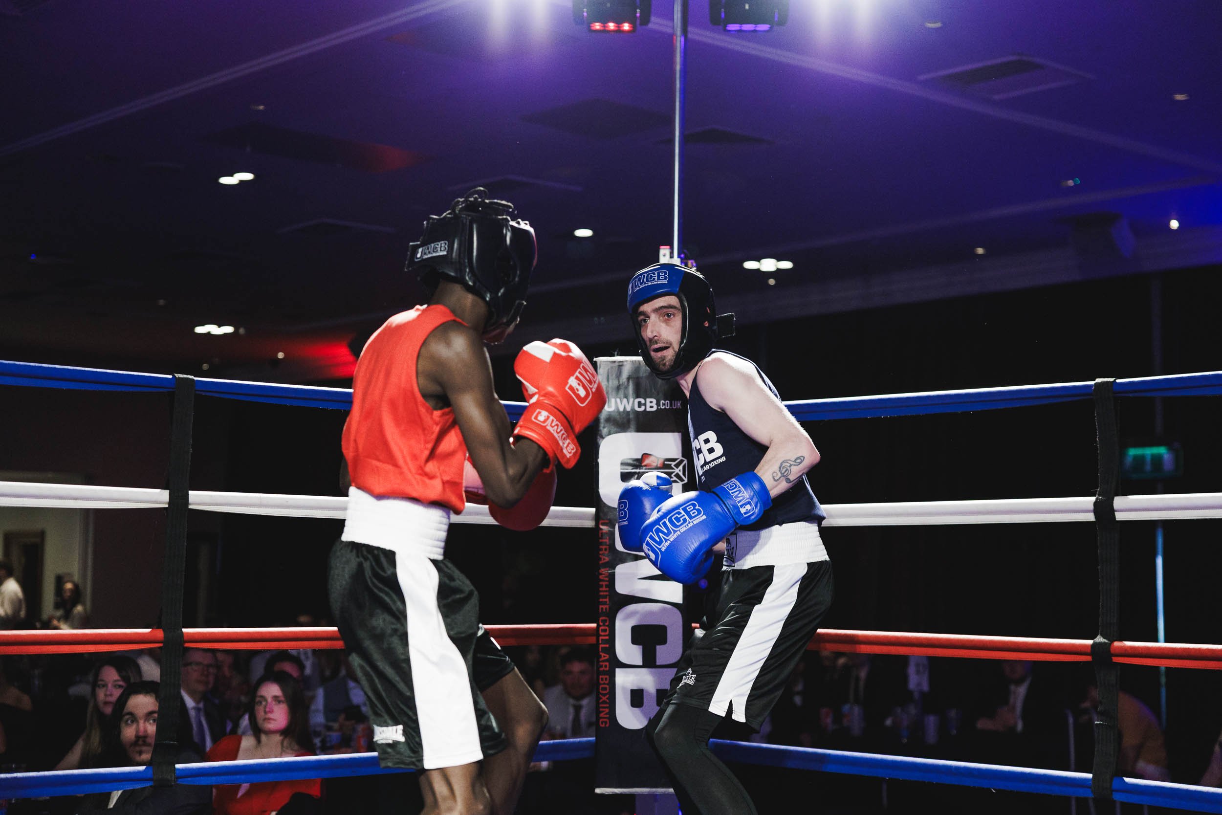 Two boxers in a ring, one in red gloves and the other in blue gloves, engaged in a match, with spectators watching in the background.