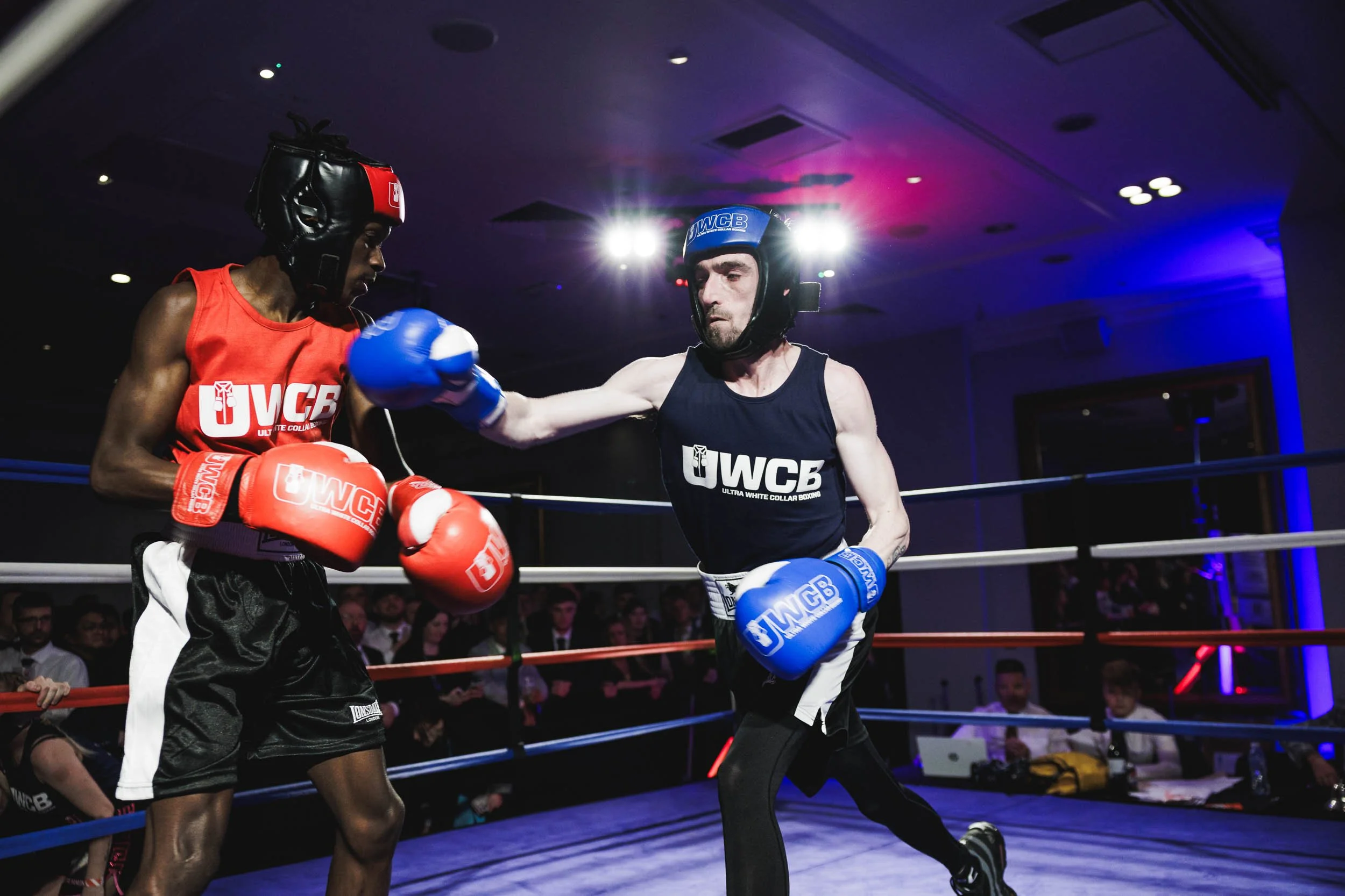 Two male boxers with protective gear sparring in a boxing ring during a match, with an audience watching in the background.