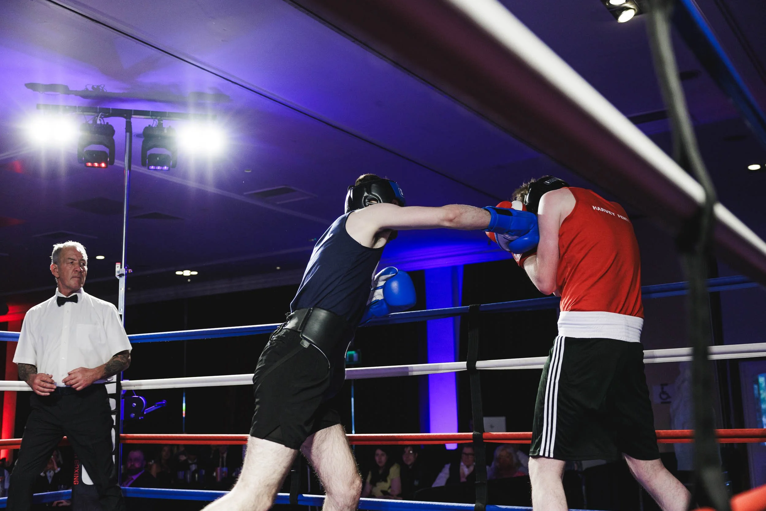 Boxers in a match, one delivering a punch while the other blocks, with a referee observing in a boxing ring with bright overhead lights and an audience in the background.
