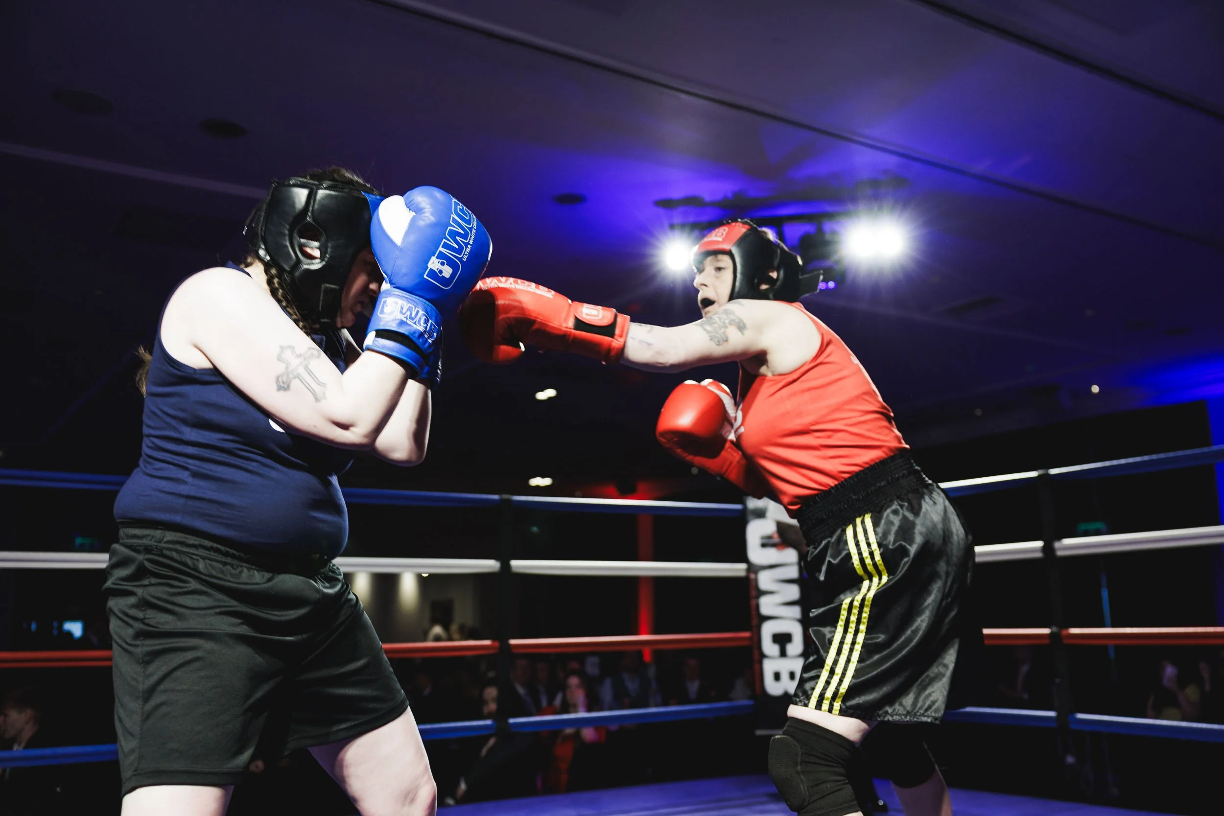 Two women wearing boxing gloves and helmets sparring in a boxing ring, with one woman throwing a punch at the other.